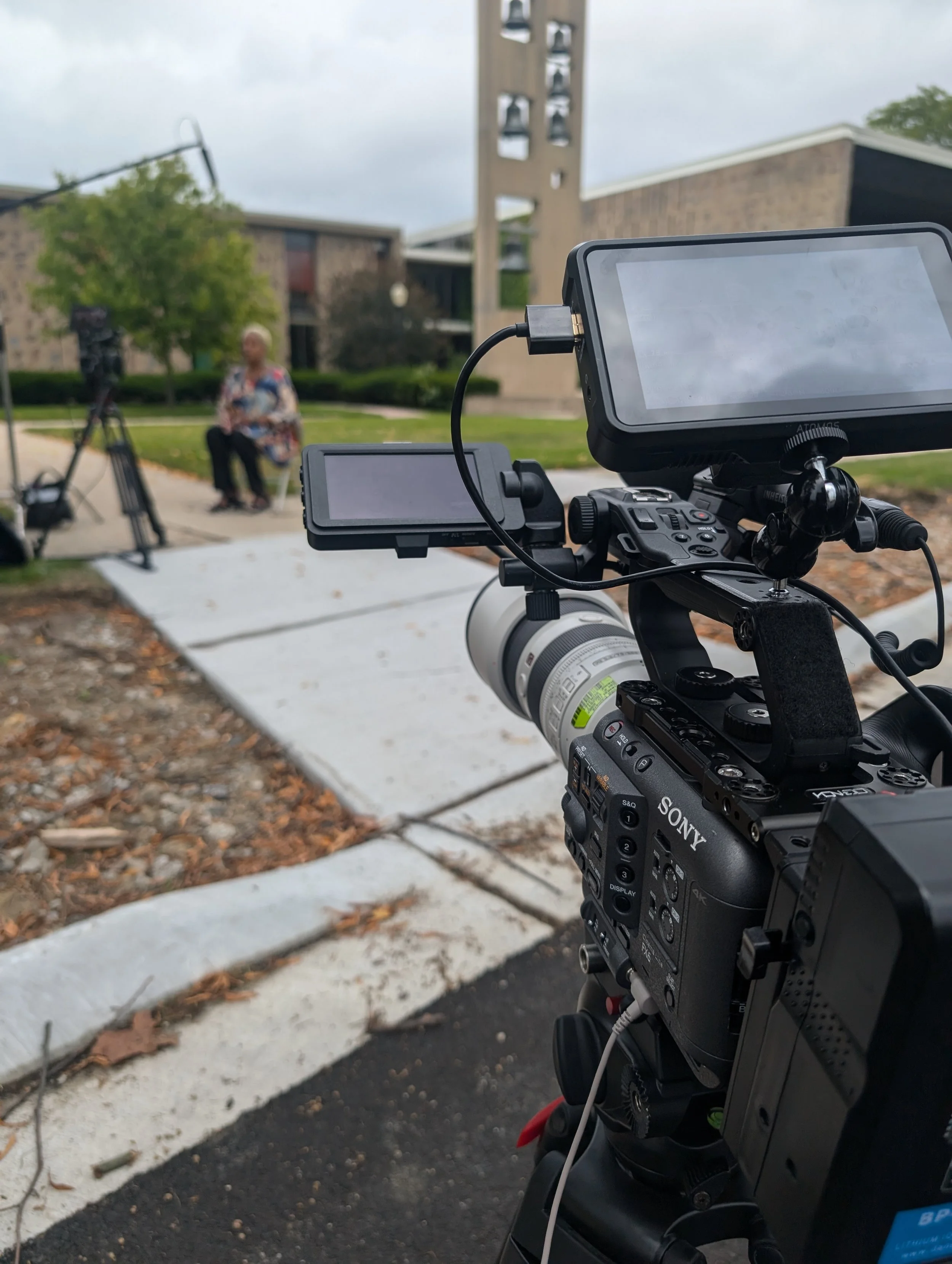A professional video camera is recording a woman sitting outdoors on a chair with a microphone and camera equipment in view, with a suburban building, trees, and cloudy sky in the background.