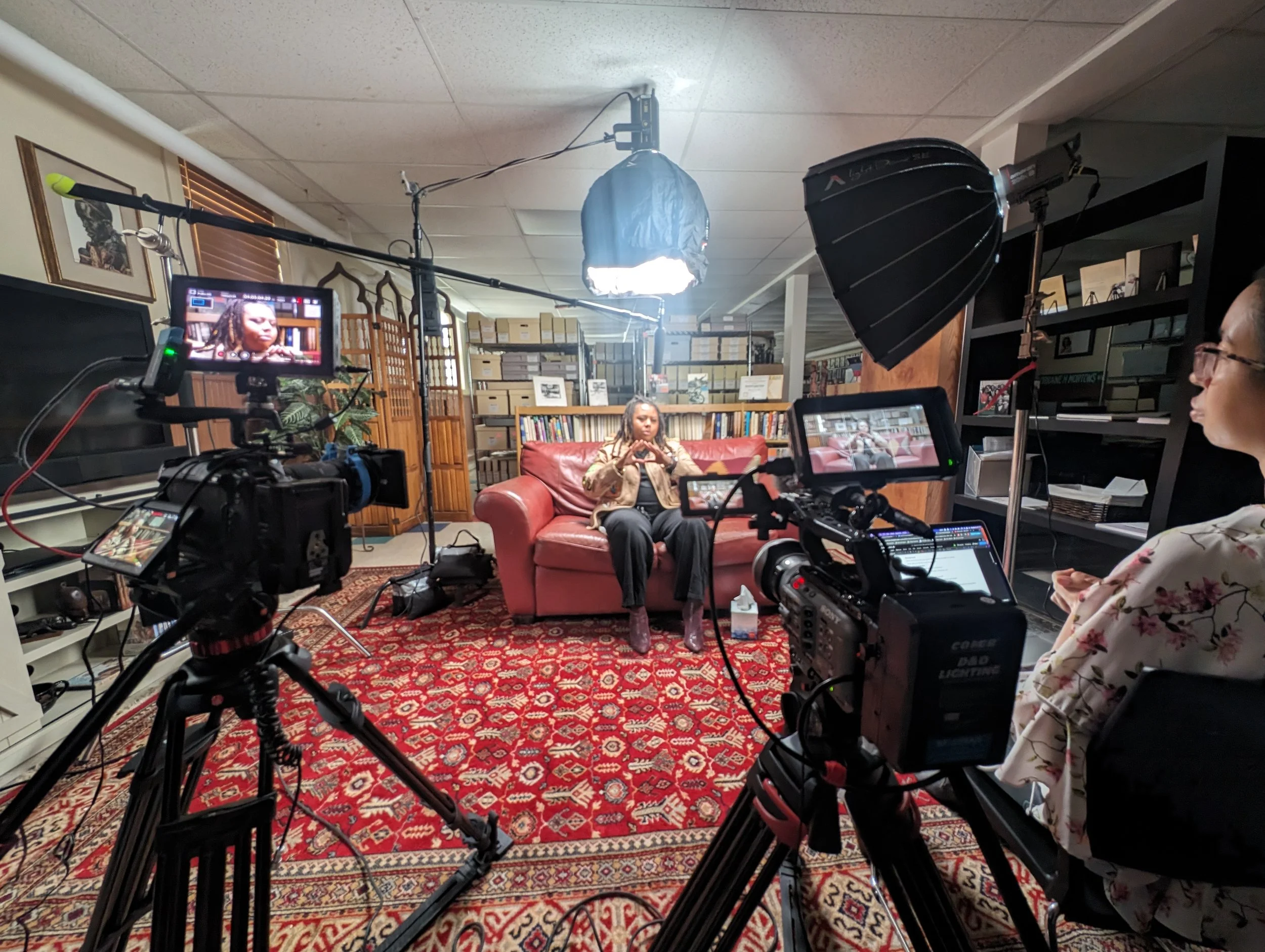 A woman with braided hair sitting on a red sofa being filmed during an interview or a video recording in a cozy book-lined room with two cameras, lighting equipment, and crew members.