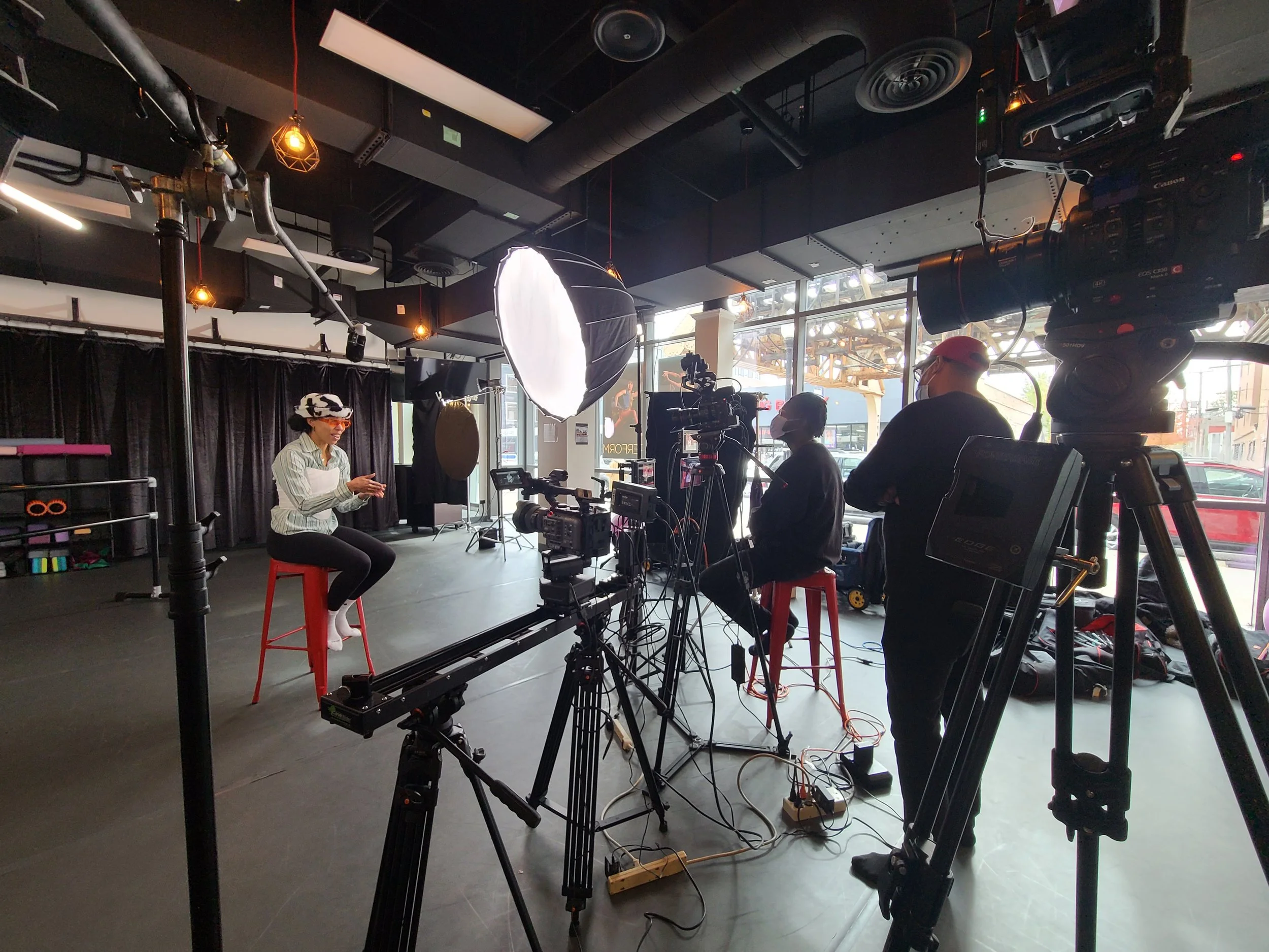 A woman being filmed in a studio with cameras and crew members, seated on a red stool, wearing a striped shirt and animal print hat.
