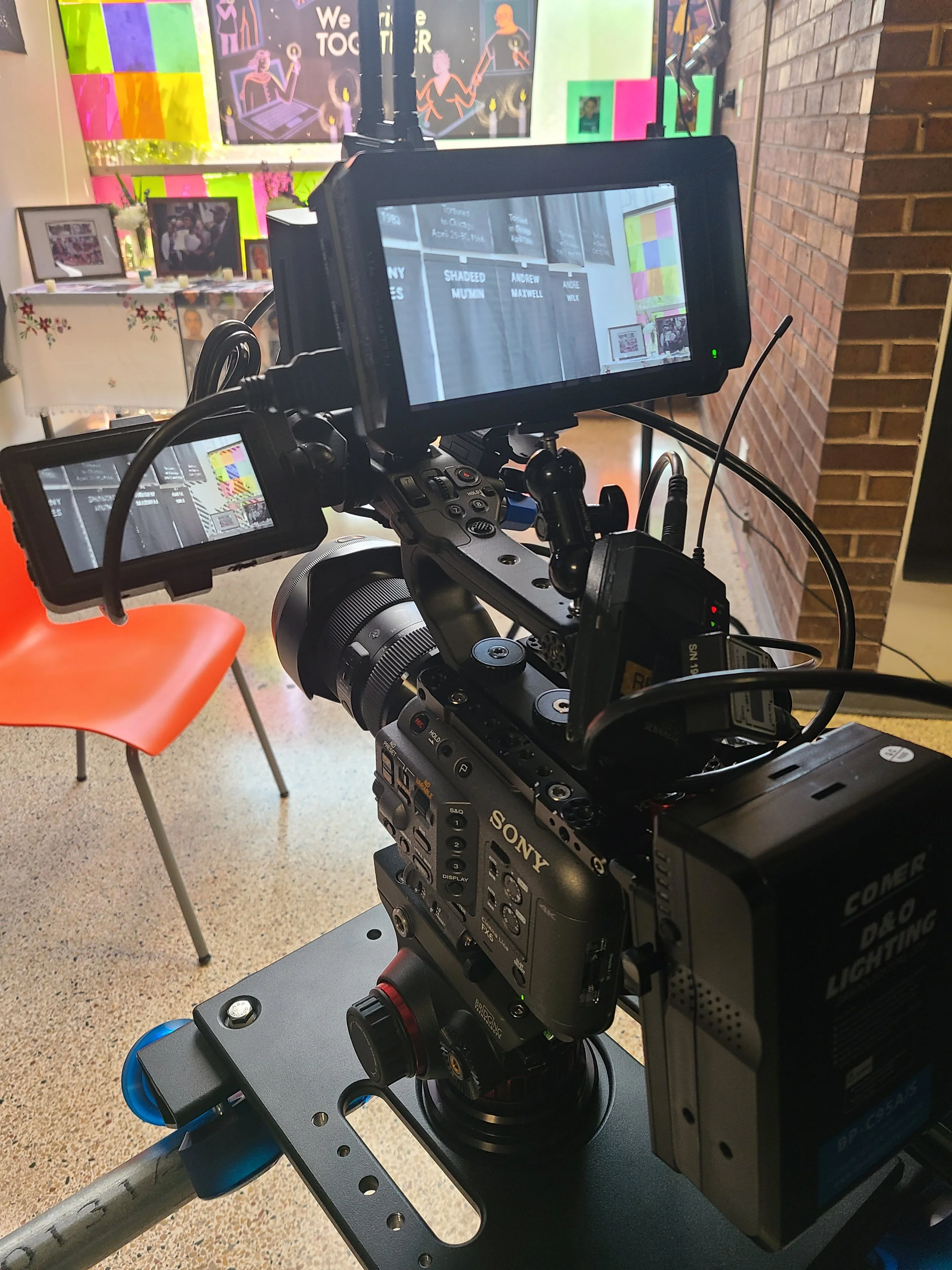 Close-up of a professional video camera setup on a tripod, recording a stage with a colorful background that includes bright, rainbow-colored posters and framed photographs on a white table. The camera screens display the stage and background.