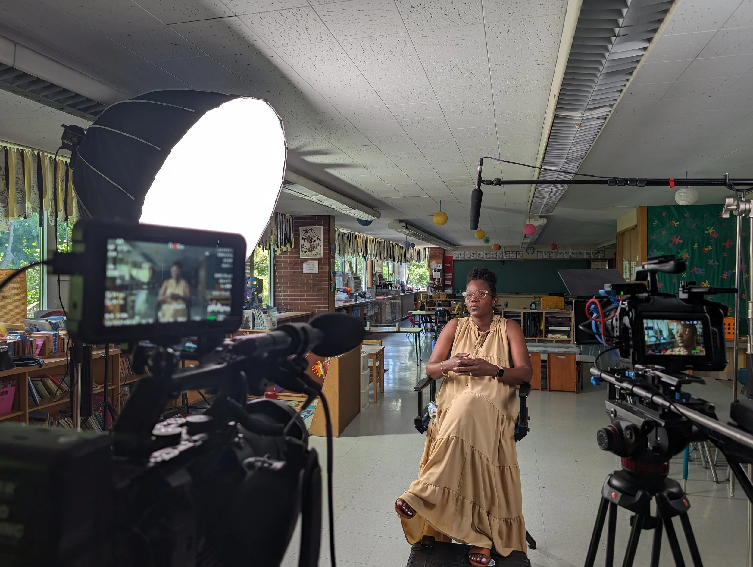 A woman in a beige dress sits in front of a camera setup in a classroom, being filmed during an interview or recording session. The classroom has bookshelves, windows, and colorful paper lanterns hanging from the ceiling.