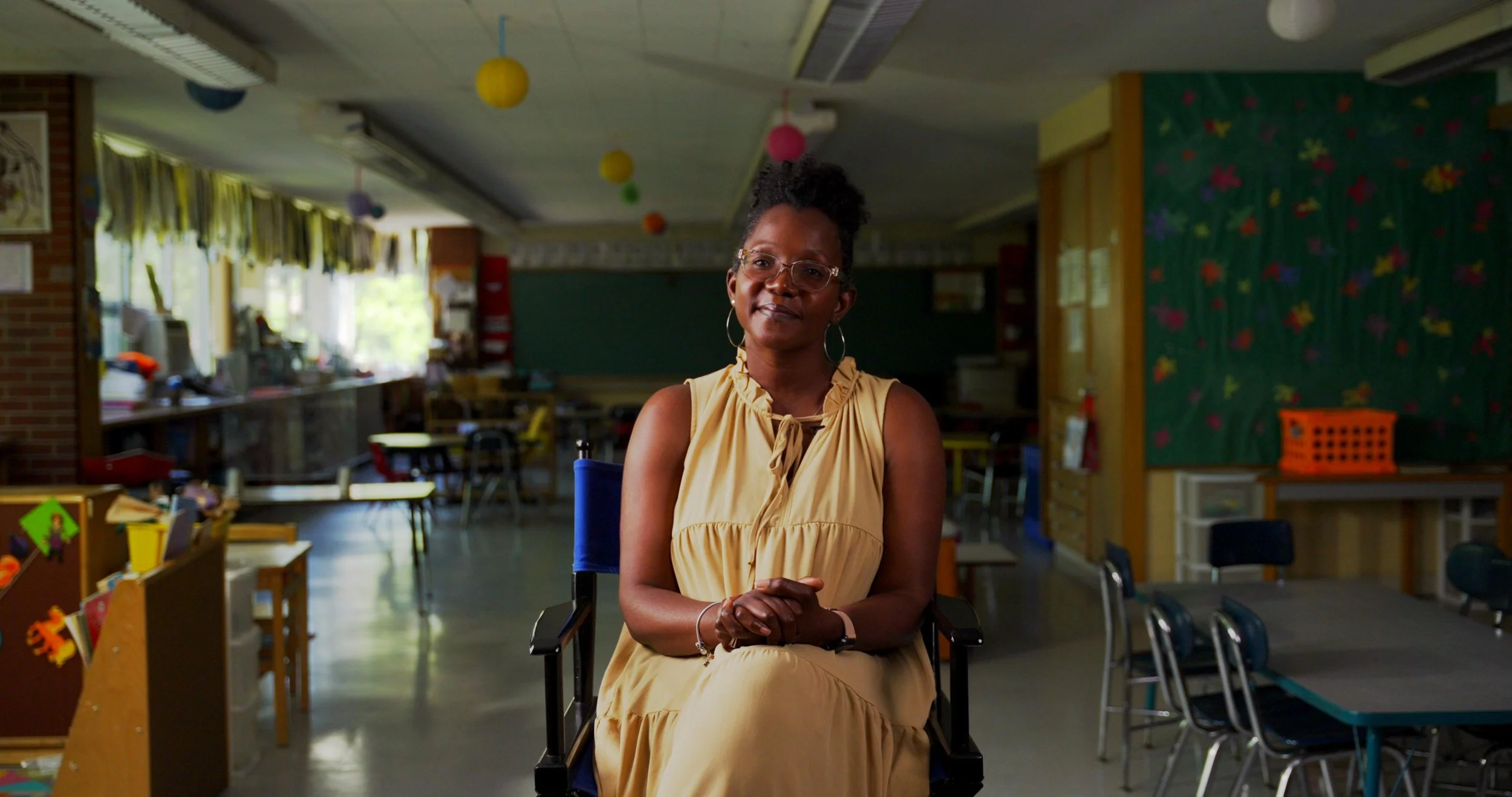 A woman sitting in a chair in a colorful classroom, with chairs, tables, and decorations in the background.