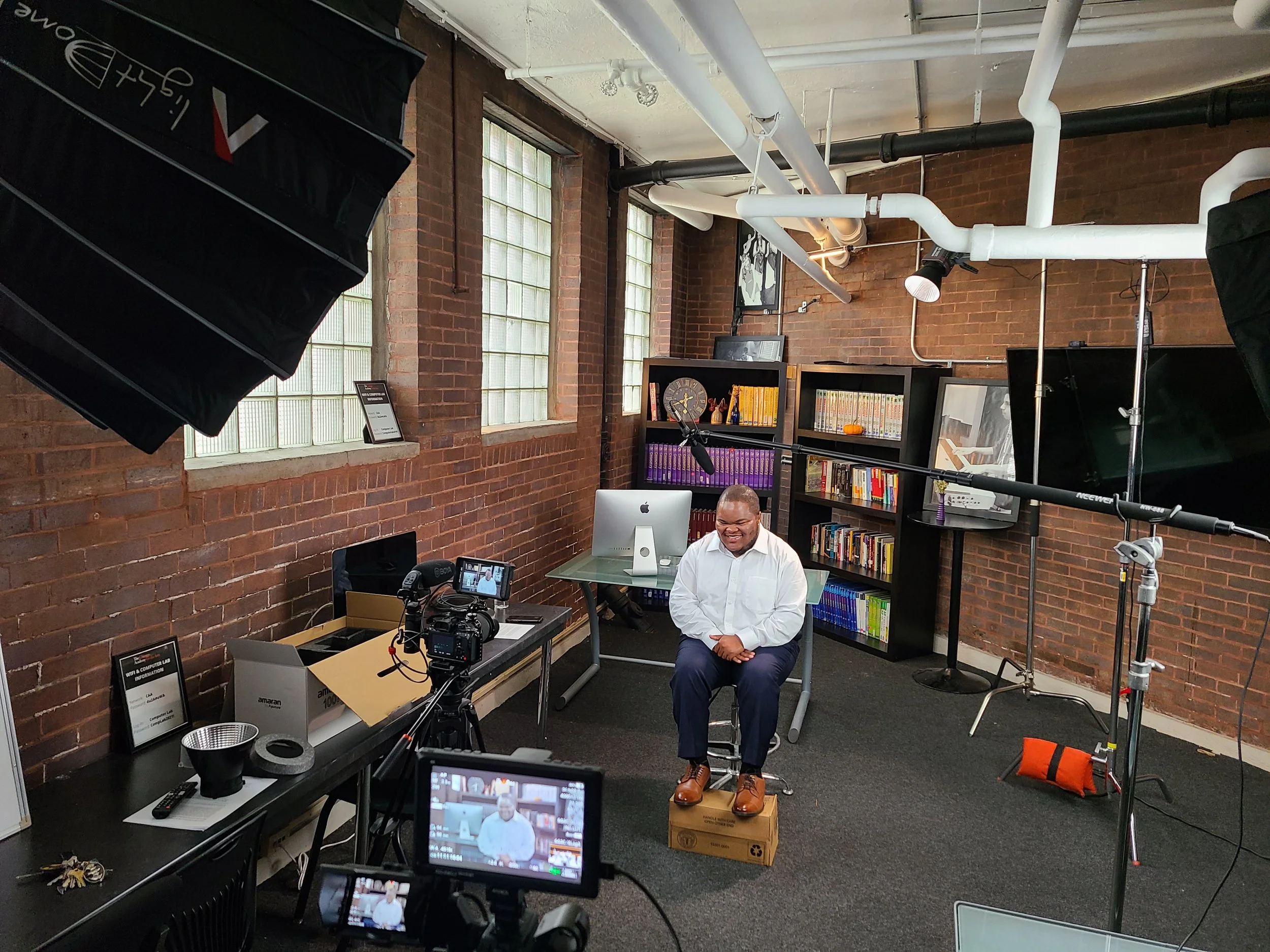 A man in a white shirt and dark pants is sitting on a cardboard box in a brick-walled room, smiling, with a bookshelf, computer, and camera equipment around him, set up for a video recording or interview.