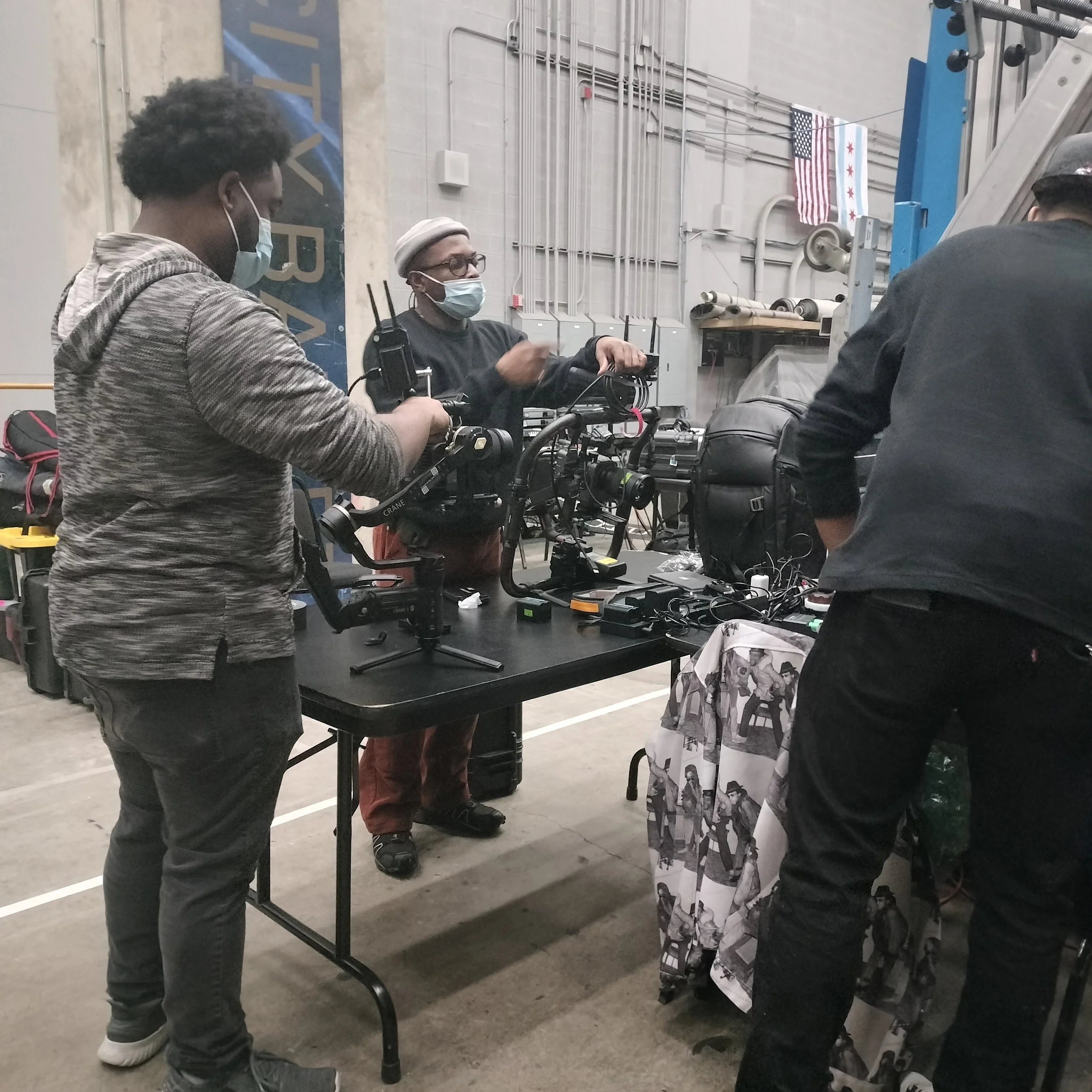 Three people wearing masks working on filming or photography equipment at a table in an industrial space, with electrical panels and American flags in the background.