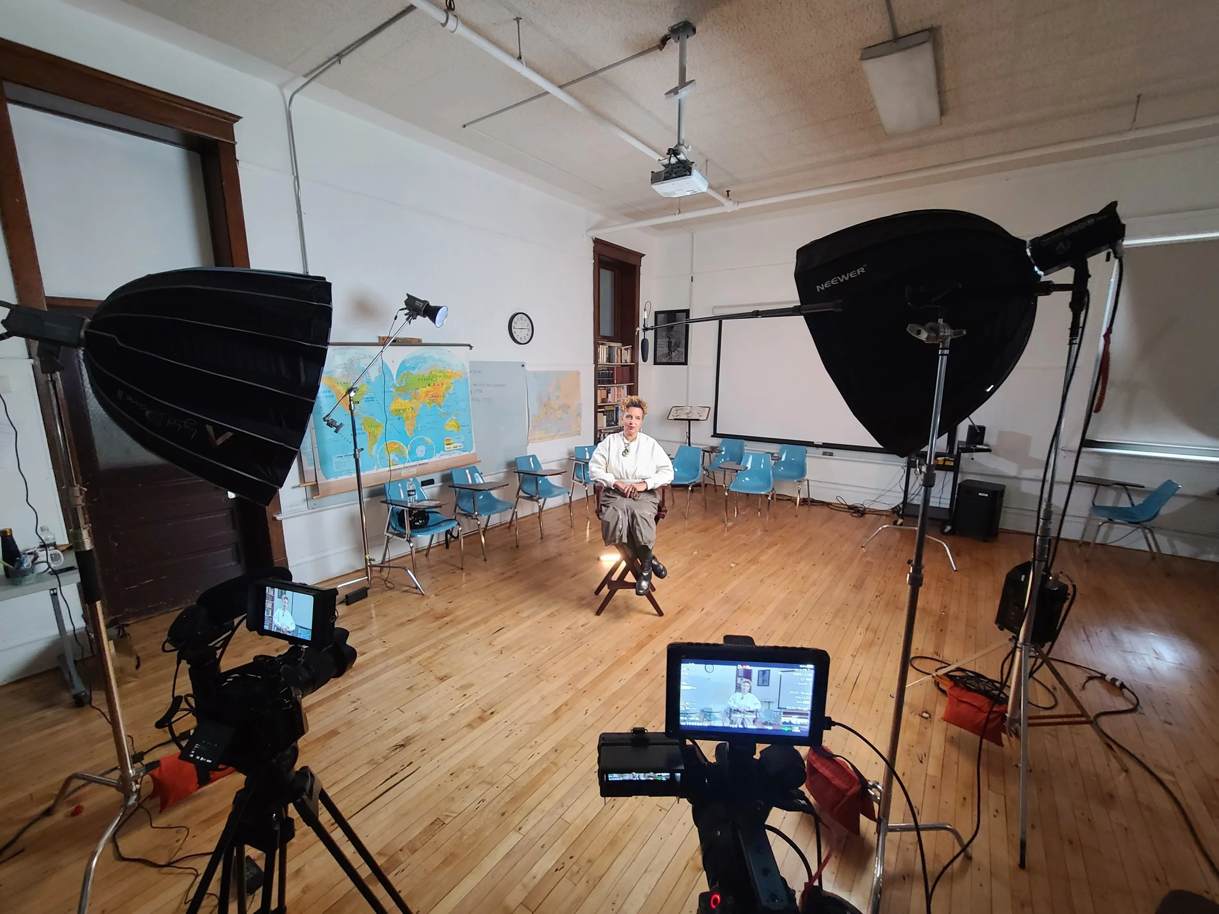 A person sitting on a stool in a classroom set up for filming or photography, with studio lights and cameras directed at them. The classroom has maps and chairs along the walls.