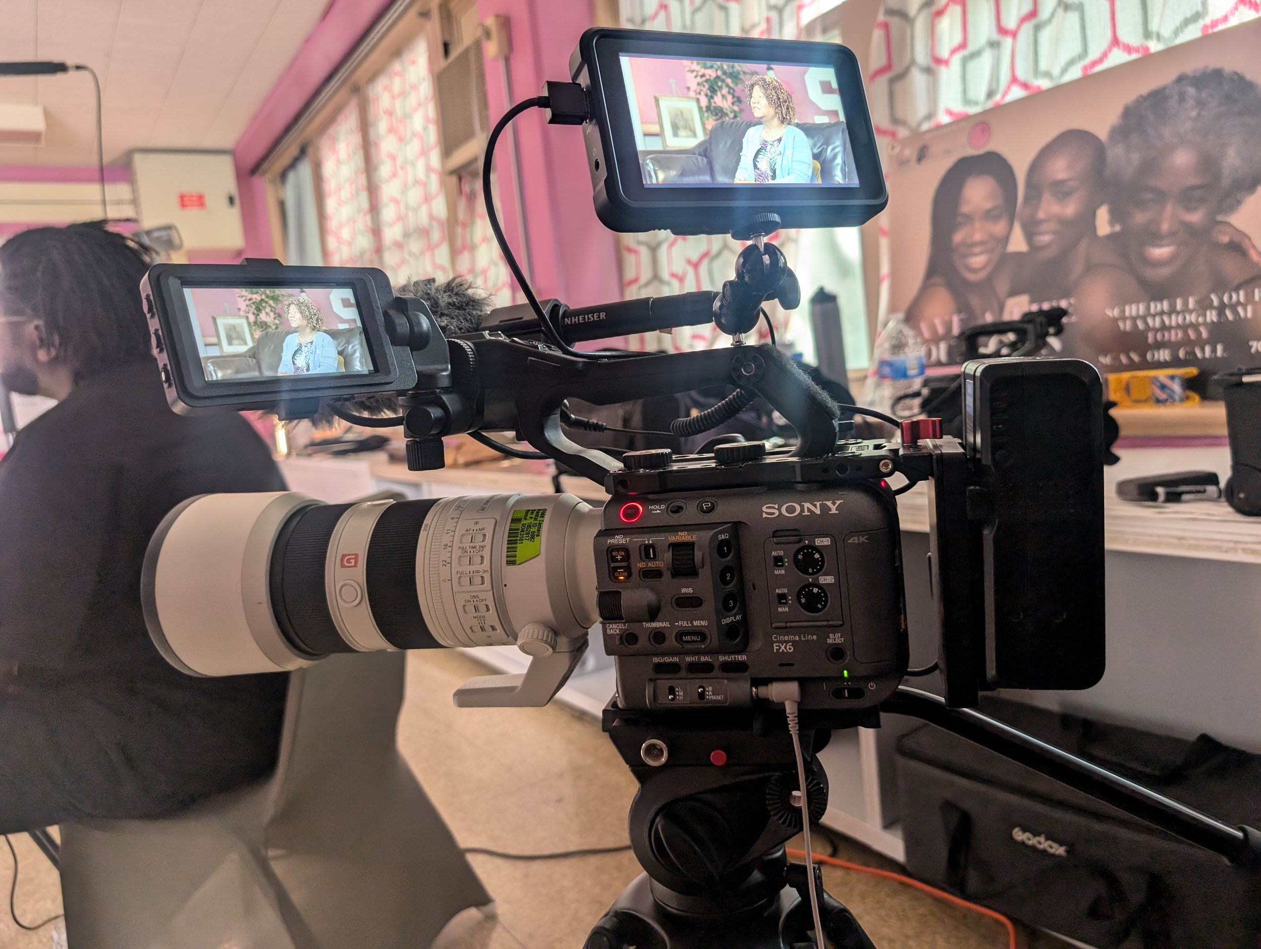 Close-up of professional video camera recording a woman speaking on a talk show set with pink and patterned curtains, a large poster of smiling women, and various objects in the background.
