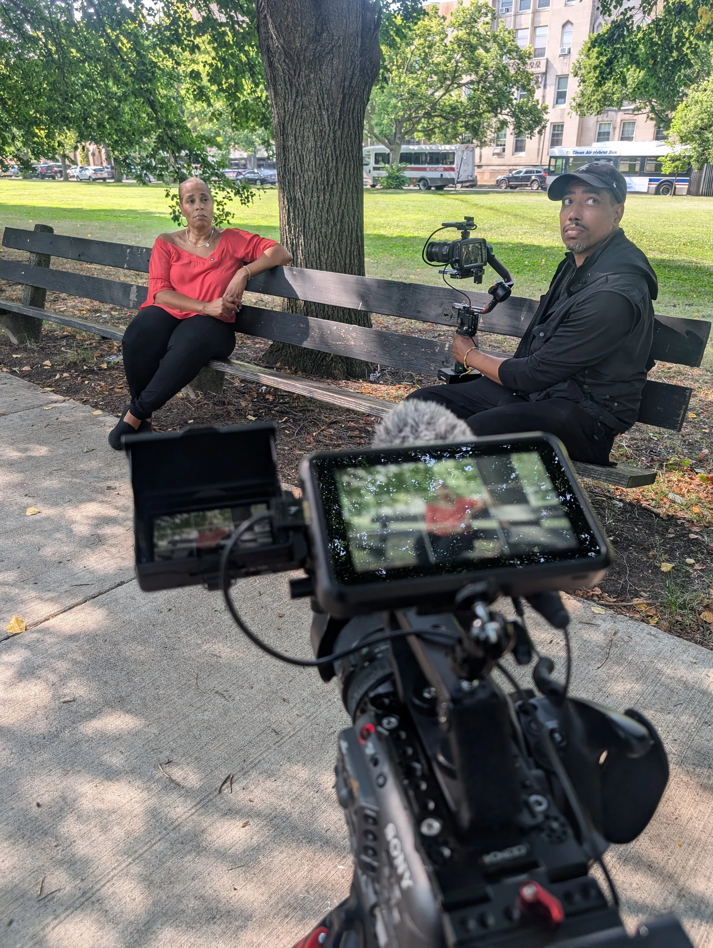 A woman sits on a park bench under a tree, with a man next to her recording her with a professional camera mounted on a stabilizer, and a camera is visible in the foreground.