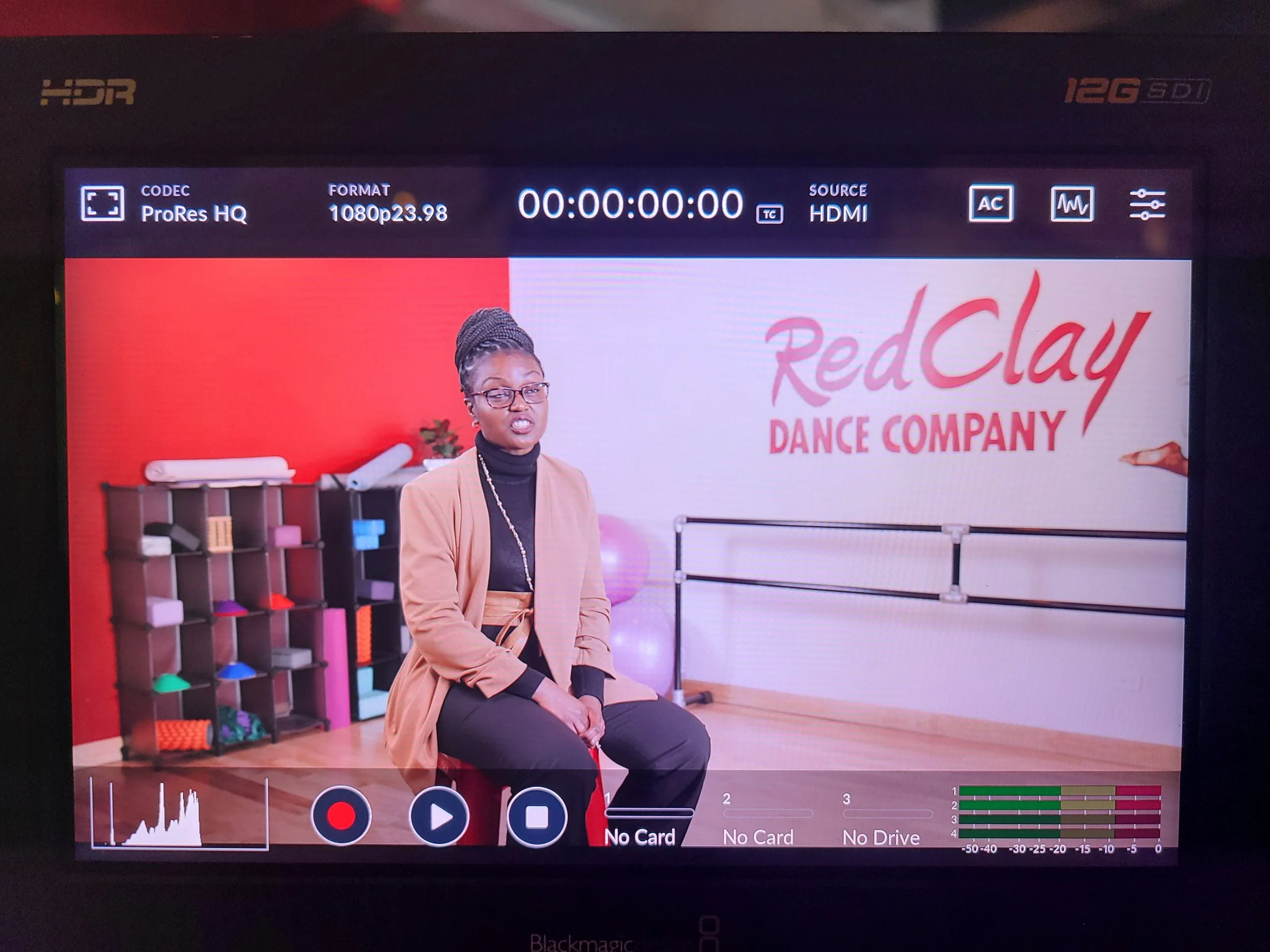 A woman sitting in a dance studio with a sign that reads 'Red Clay Dance Company' in the background. There are fitness mats on the shelf and ballet barres on the wall.