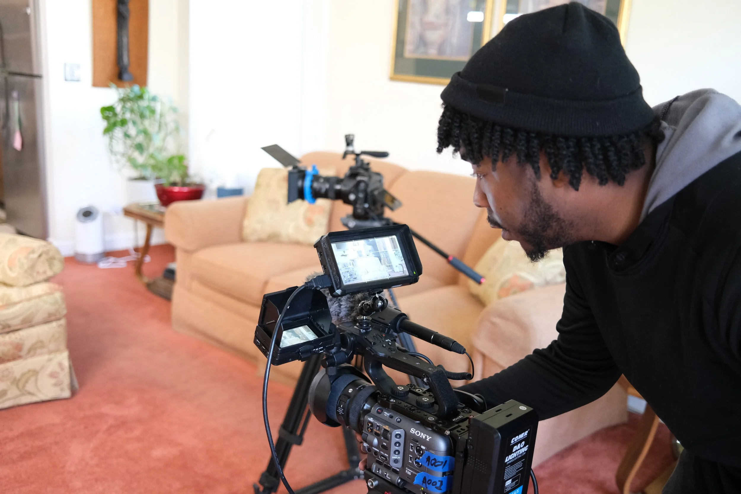 A person is setting up a professional video camera on a tripod in a living room, with another camera mounted on a stabilizer in the background.