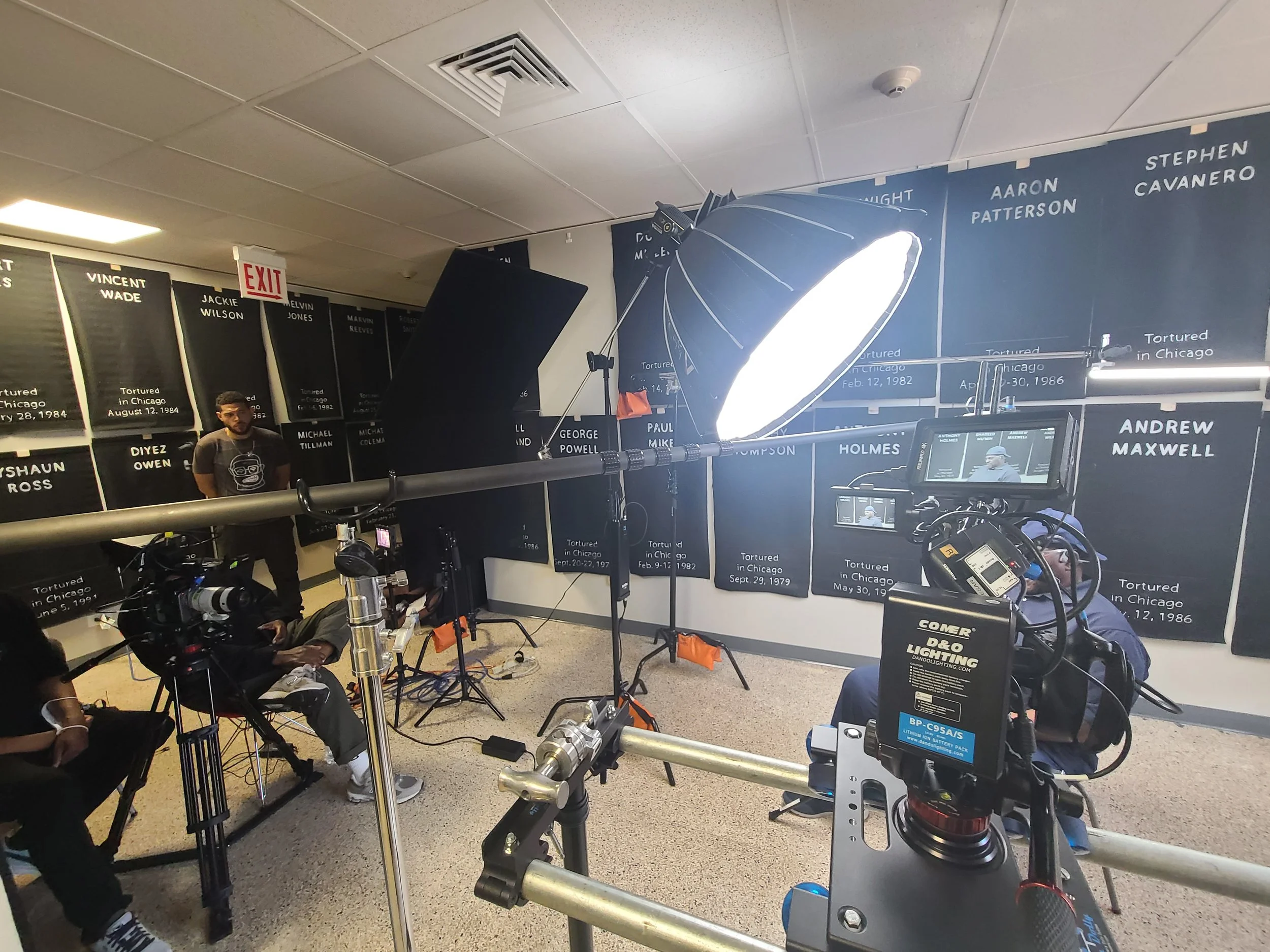 Behind the scenes of a film or documentary shoot, with cameras, lighting equipment, crew members, and a backdrop with names and dates related to torture in Chicago.
