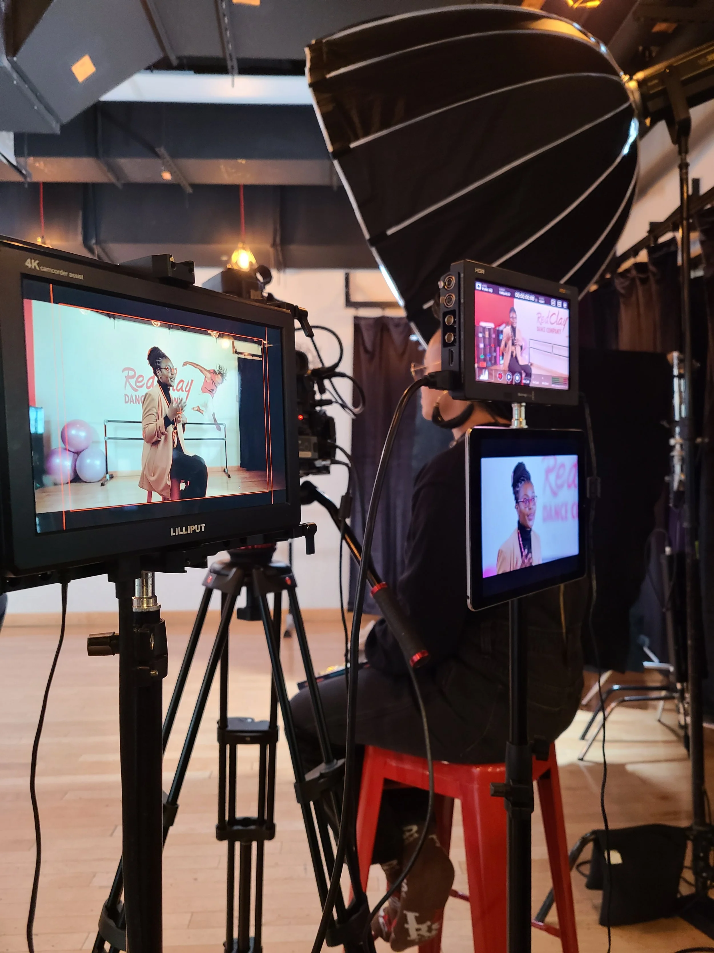 A woman is seated on a stage in a dance studio, holding a microphone, with balloons and a sign that reads 'Rea Clay Dance Studio' in the background. The scene is being recorded or photographed with multiple cameras and monitors capturing her from dif