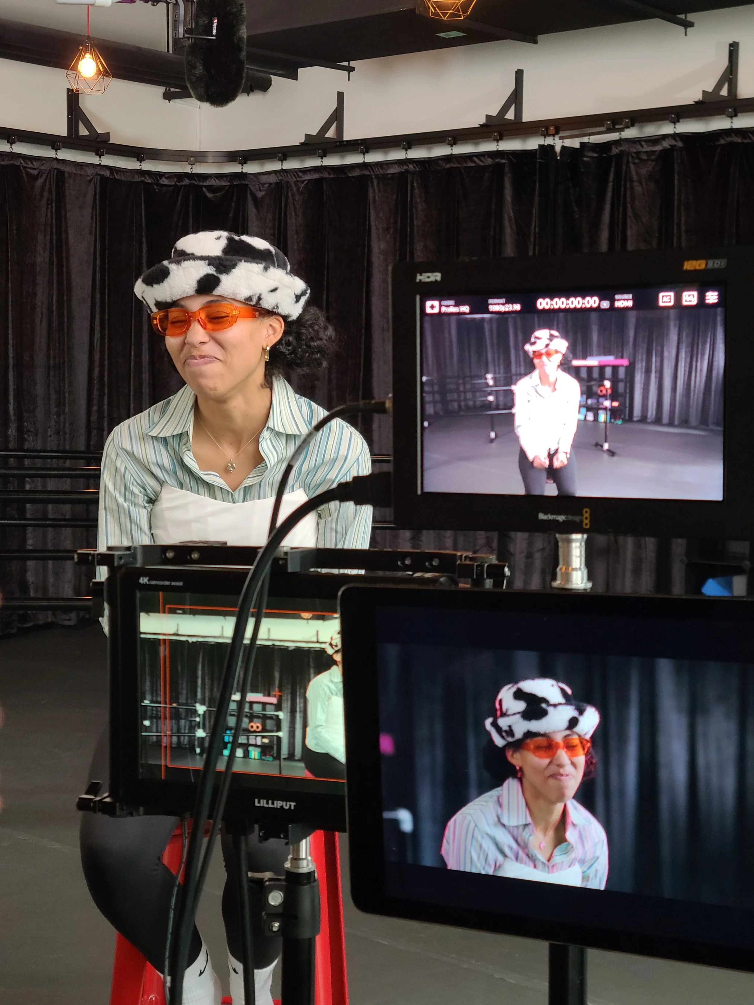 A woman with curly hair, wearing orange sunglasses, a striped shirt, and a black-and-white spotted hat, standing in front of a black curtain with video screens around her showing her image.