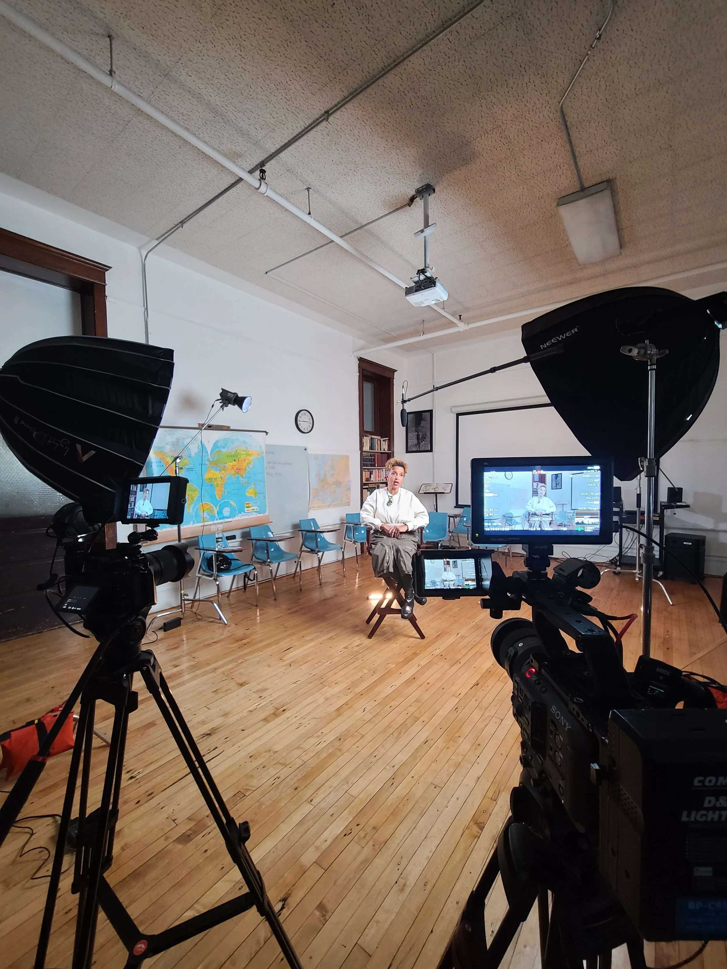 A woman sitting on a chair in a recording studio or classroom setup, surrounded by professional video camera equipment, softboxes, a monitor, a world map, and a whiteboard, with chairs and bookshelves in the background.