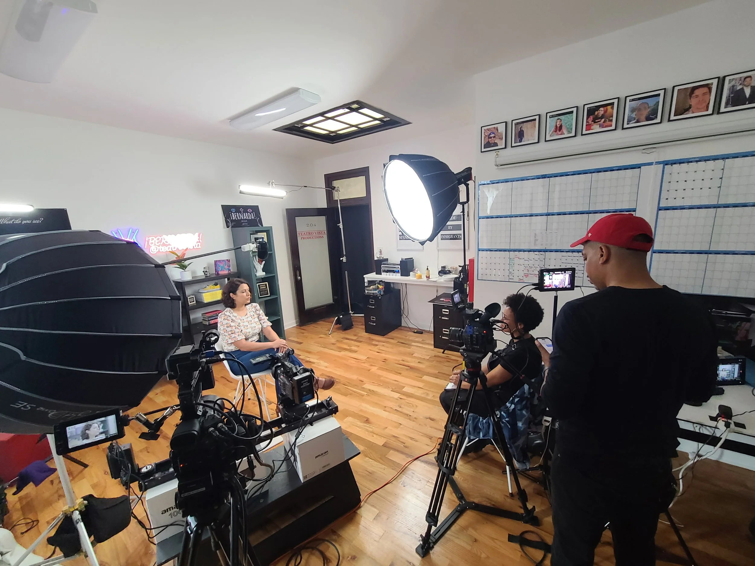Filming crew recording a woman in a colorful blouse sitting on a chair in a room with wooden floors and white walls with framed photos. Equipment includes cameras, lights, and a large umbrella reflector.