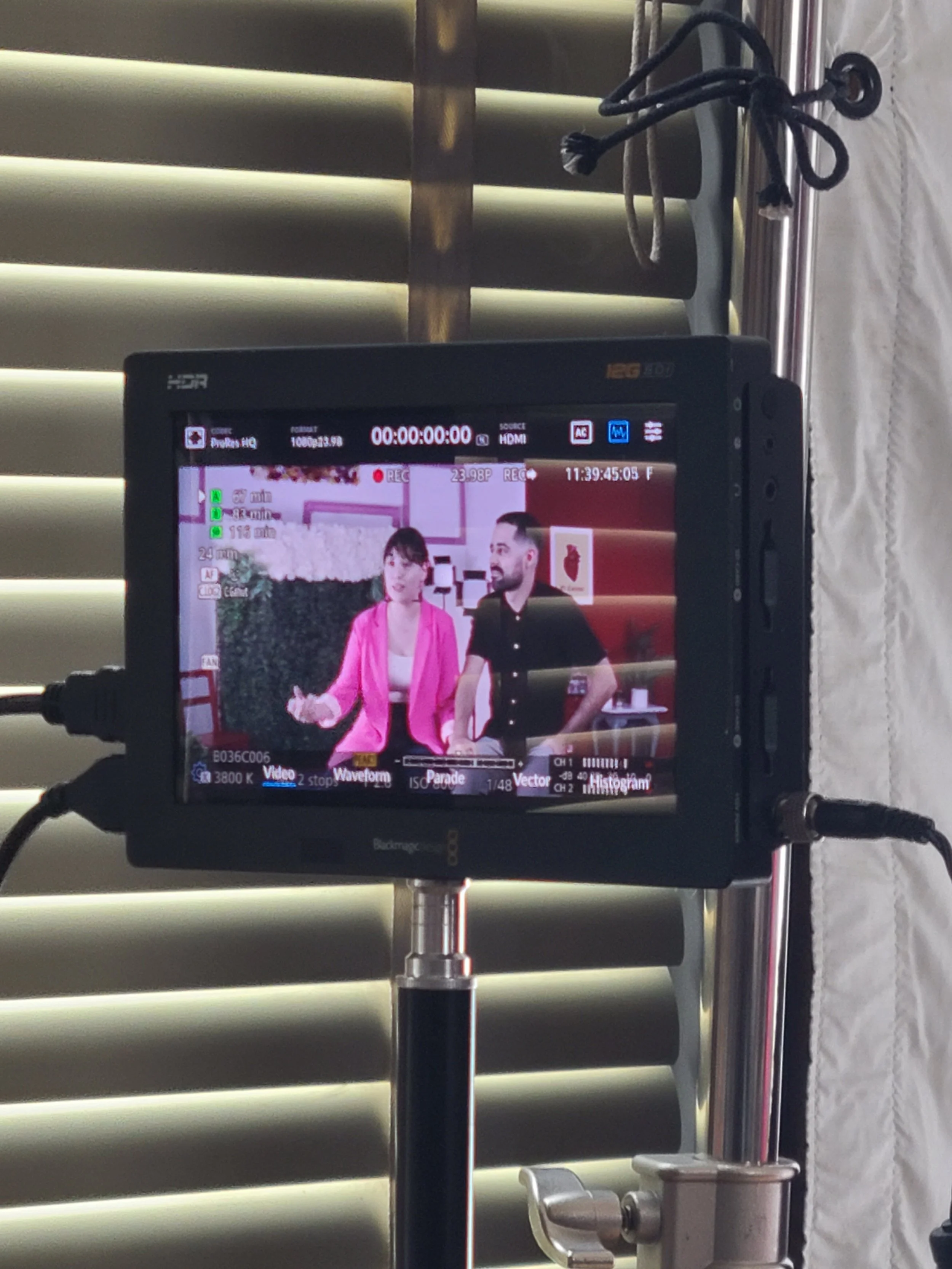 A camera monitor shows two people sitting and talking, with a pink and red background and window blinds behind them.