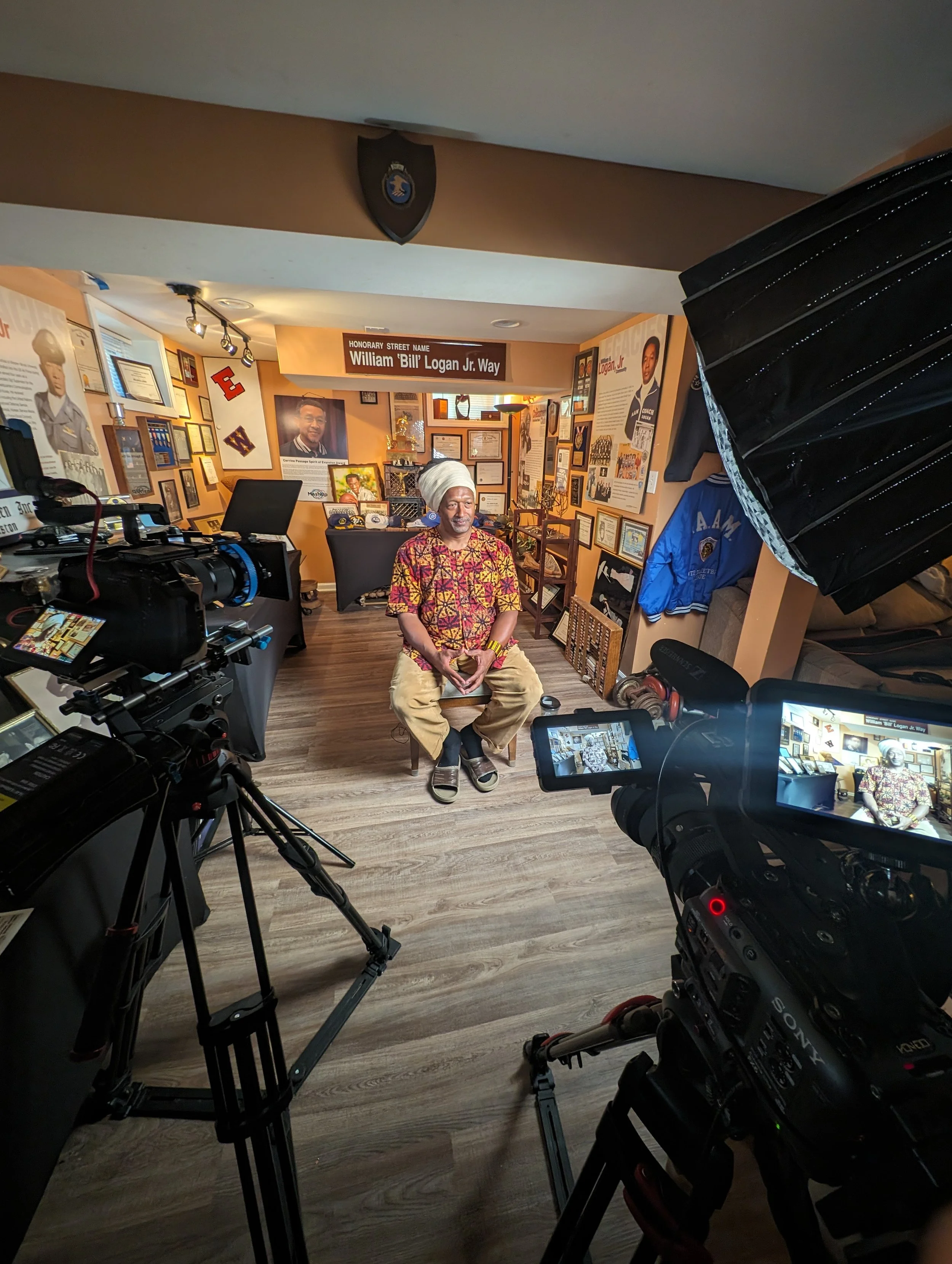 A man sitting on a chair in a room filled with framed photographs and certificates, with professional video camera equipment capturing him for an interview or documentary.