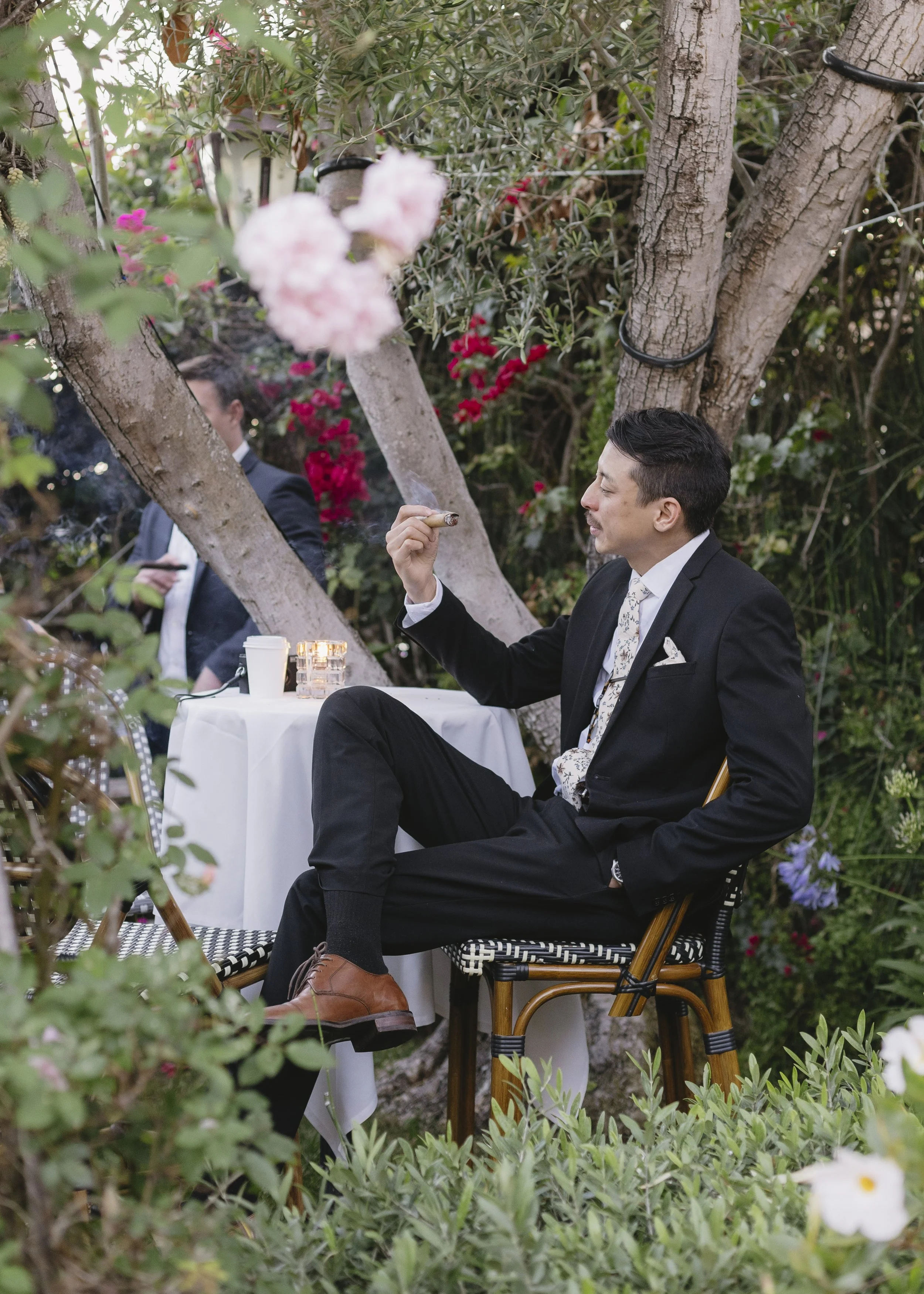 A man in a black suit and tie is sitting outdoors at a table at the Guiseppe's event venue in Pismo Beach Califorina, smoking a cigar. The setting is lush and garden-like, with trees, bushes, and flowers around him.