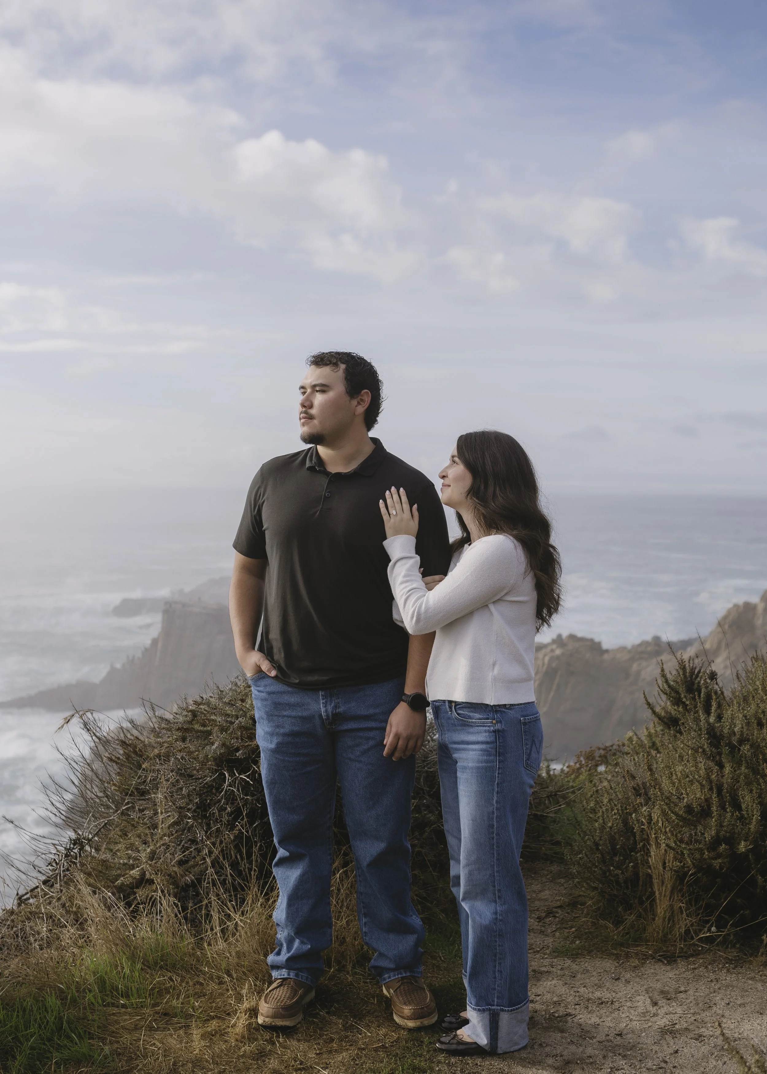A man and woman standing on a cliffside overlooking the ocean with cloudy skies at Painters Point in Carmel California, the woman touching the man's shoulder and both looking away.