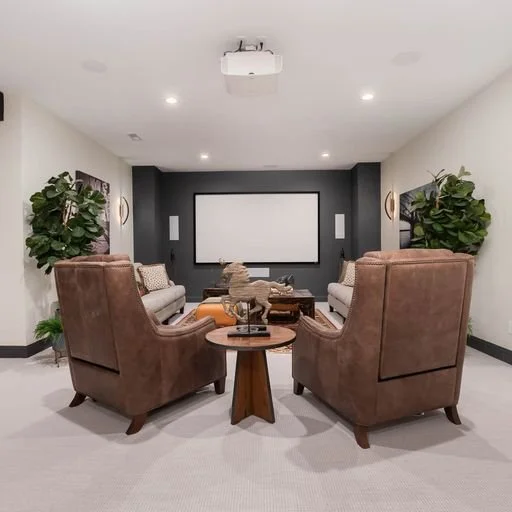 Home theater room with a large projection screen, two armchairs, two small sofas, side tables, wall-mounted speakers, and decorative plants.