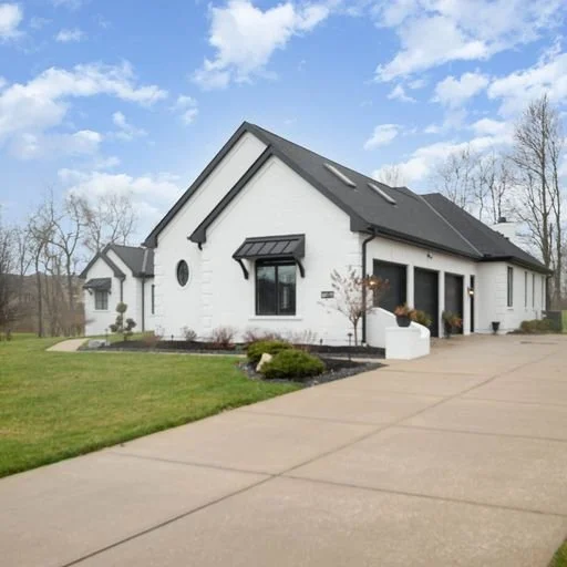 A white house with black roof and trim, with a small front lawn and a paved driveway, under a partly cloudy sky.
