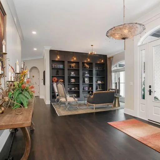Spacious living room with dark hardwood floors, a large black bookshelf, two vintage style chairs, a dark sofa, and decorative lamps, with a front door and window on the right side.
