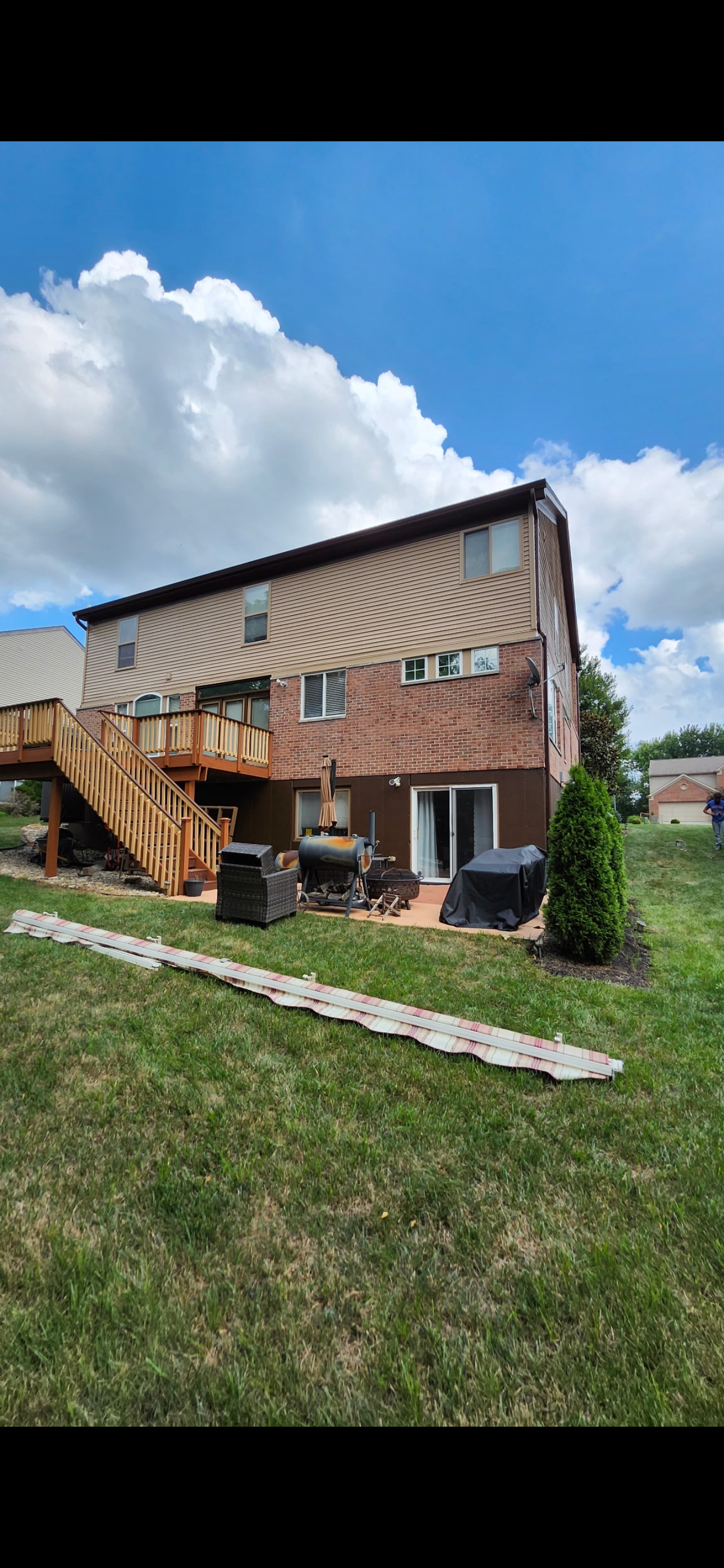 Backyard view of a multi-story house with a wooden deck and stairs, patio furniture, a barbecue grill, and a lawn with a rolled-up rug or outdoor mat, under a partly cloudy sky.