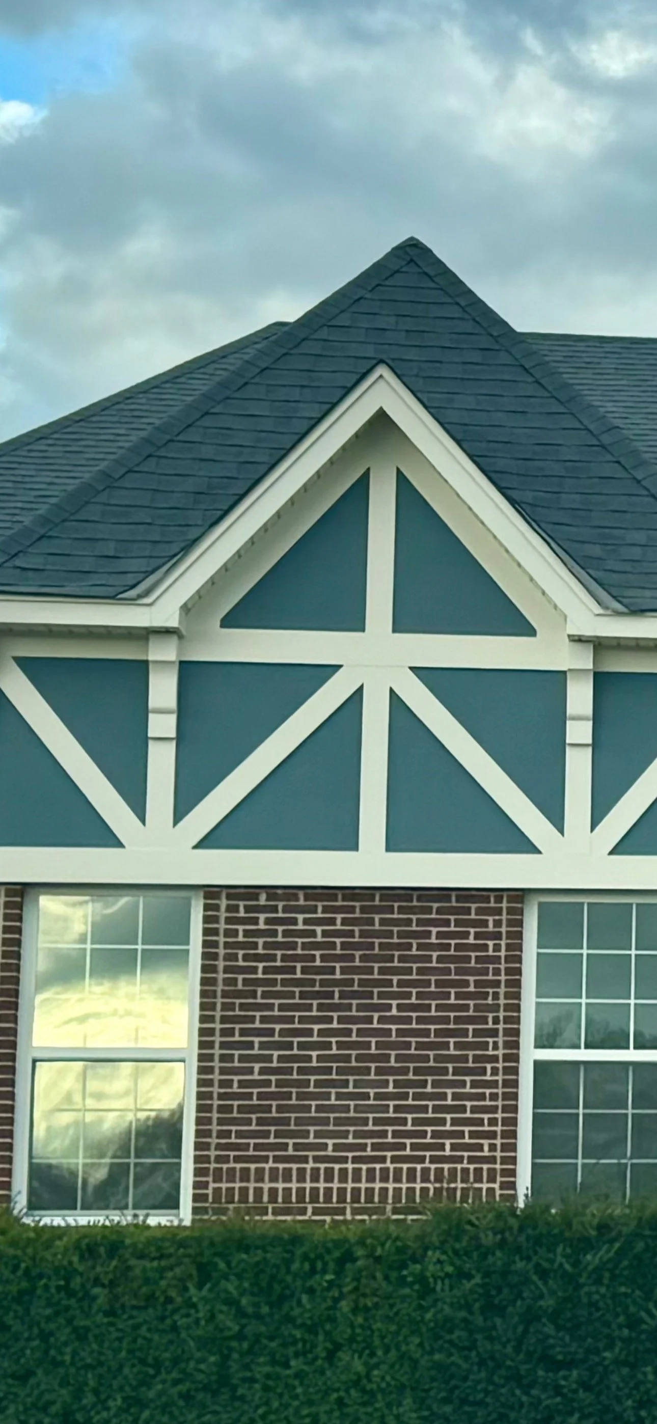 The image shows the upper part of a house with a steep dark gray shingled roof, blue and white decorative Triangular and X-shaped trim, two large windows with white framing, a brick exterior, and a well-maintained green hedge in the foreground, under
