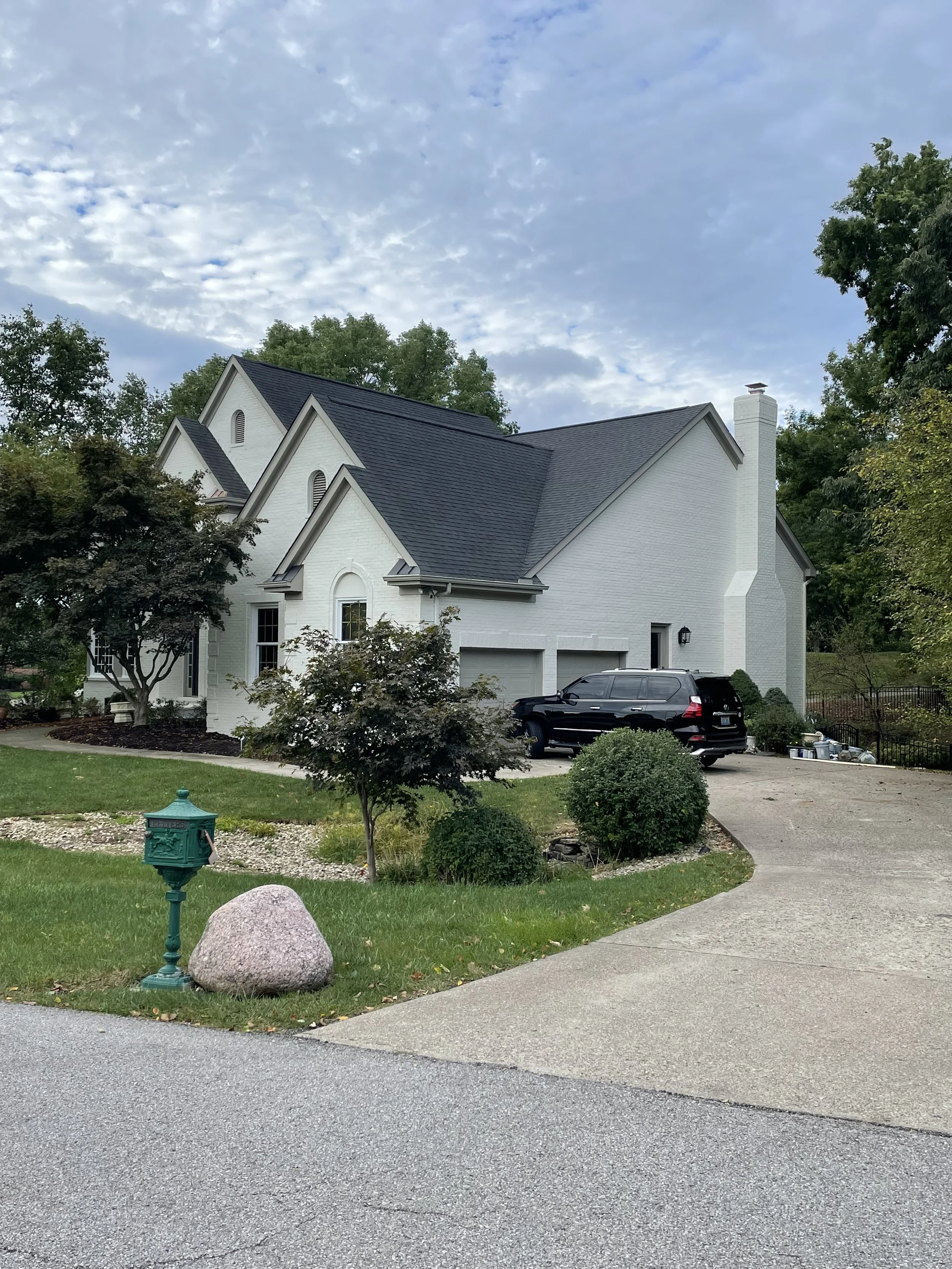 A white house with a gray roof and a chimney, surrounded by trees and shrubs, with a driveway and a parked black SUV in front.
