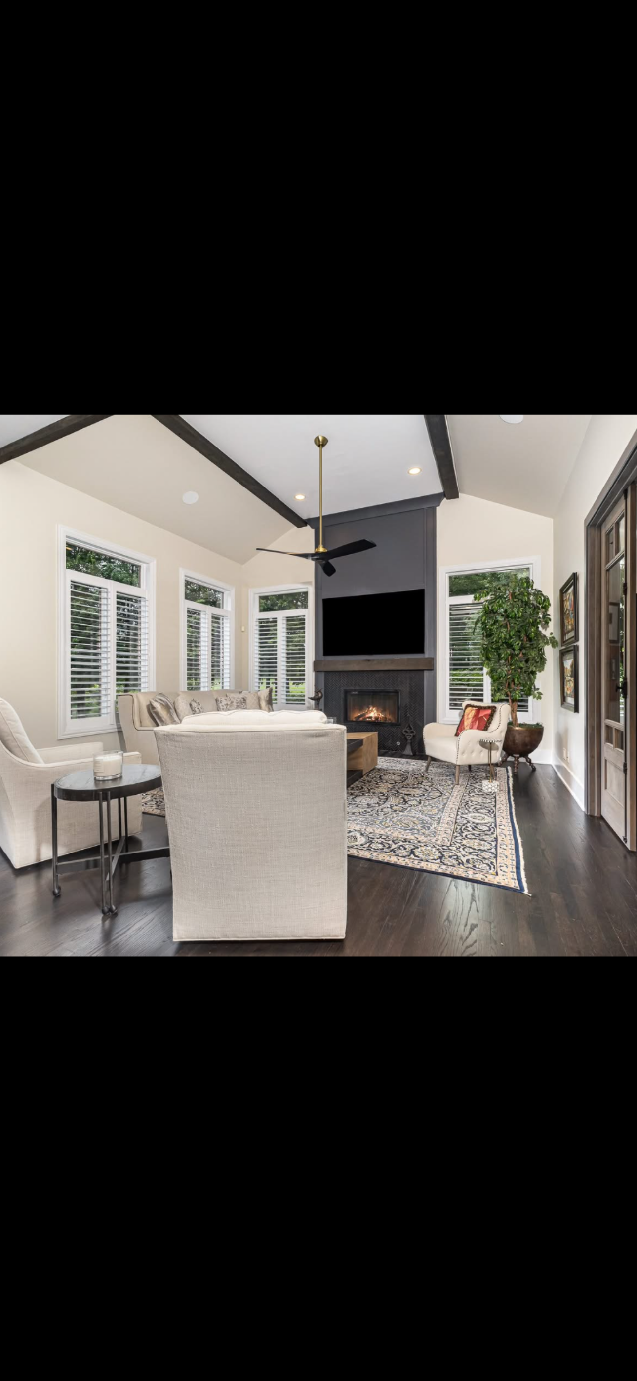 Living room with beige sofas, a patterned area rug, a fireplace, a black TV above the fireplace, large windows with white shutters, a potted plant, and wooden accents.