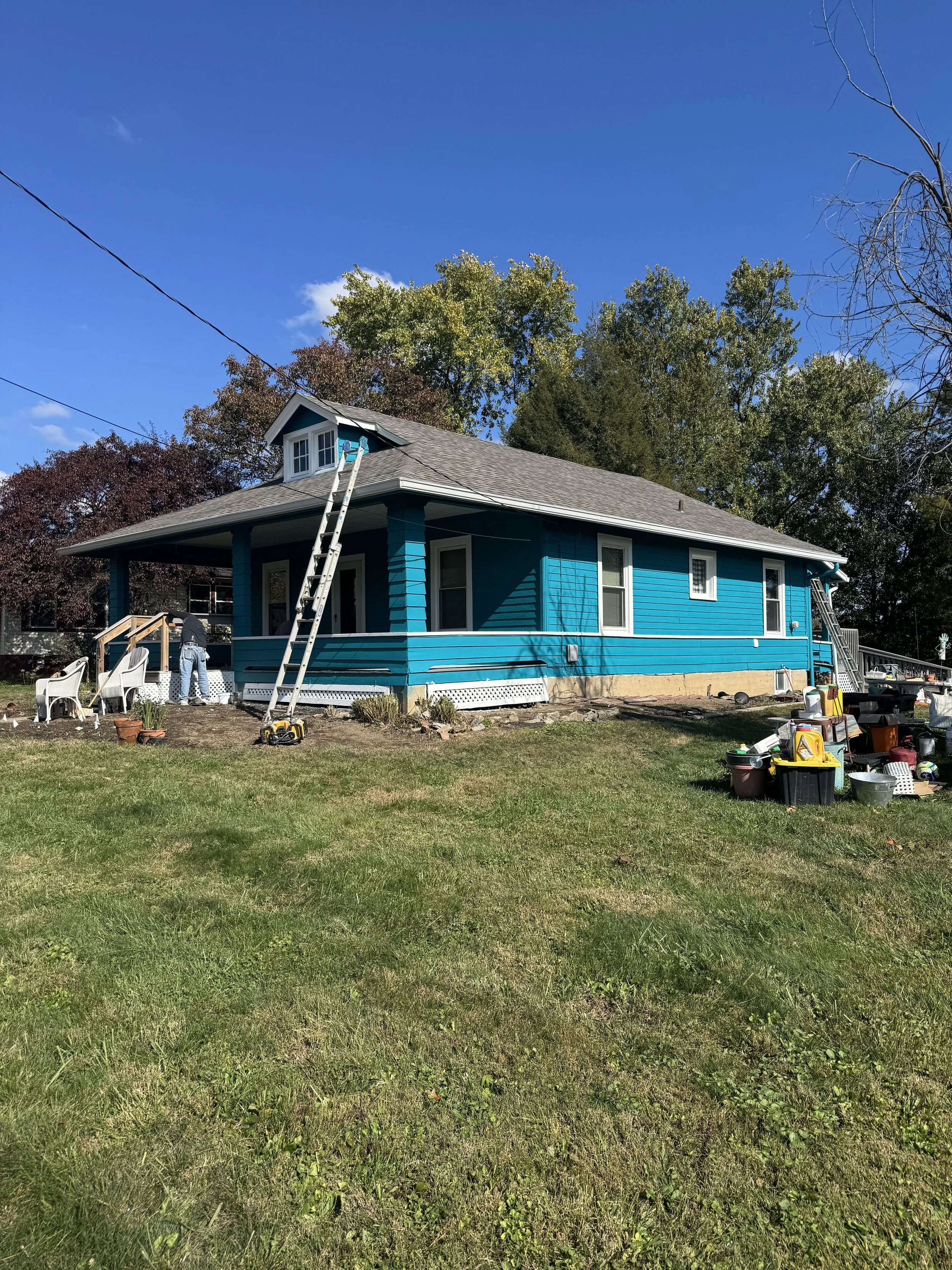 Blue house under a bright blue sky with trees in the background, ladders leaning against the house, and various tools and supplies on the ground.