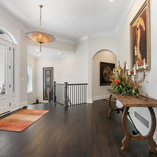Entryway and foyer of a home with a dark hardwood floor, a rug, a small table with floral arrangements, framed artwork on beige walls, a stairway with a black railing, and a chandelier hanging from the ceiling.