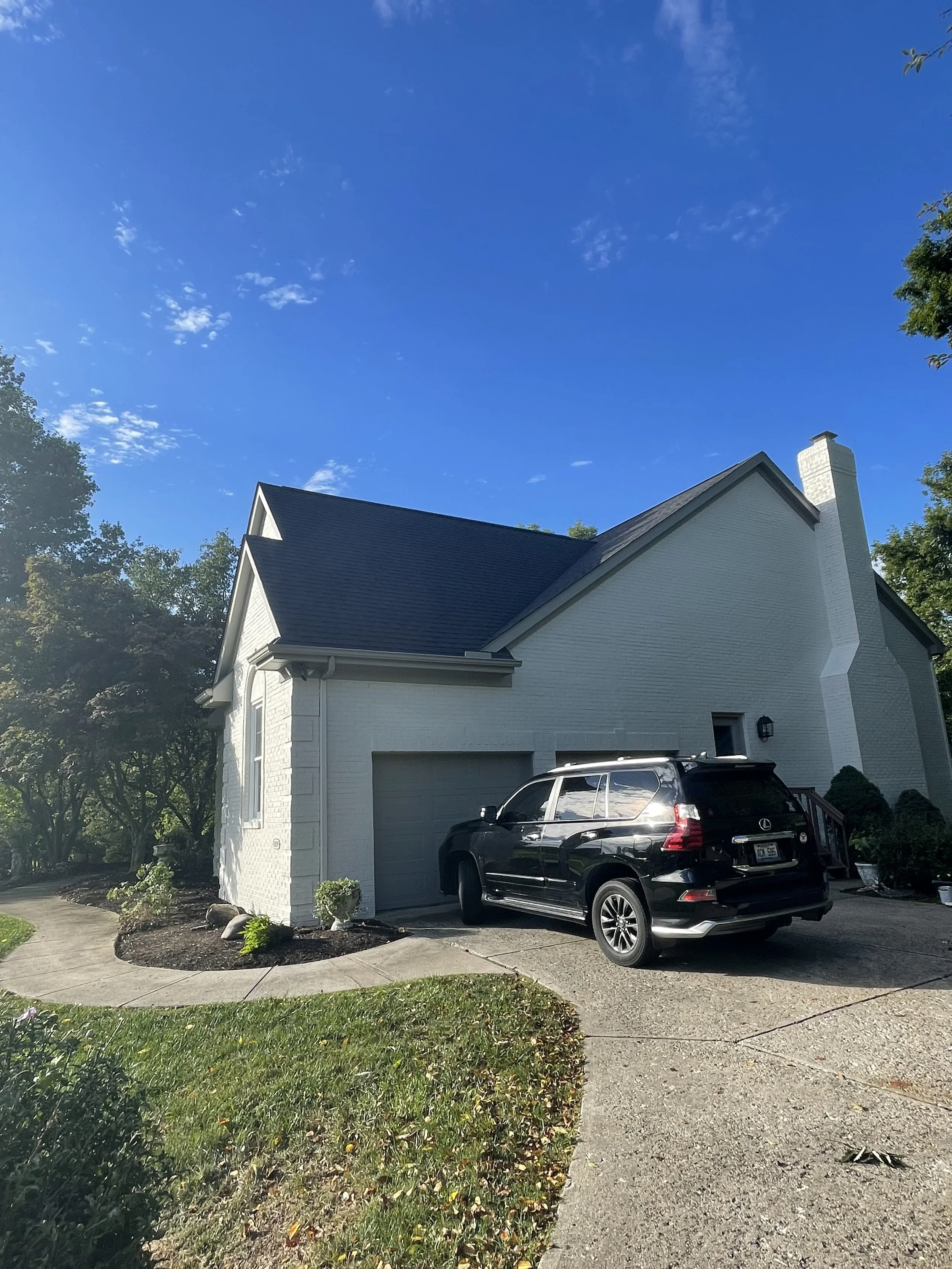 Front view of a white house with a dark gray roof, a driveway with a black SUV, and green lawn with landscaping. The sky is blue with scattered small clouds.