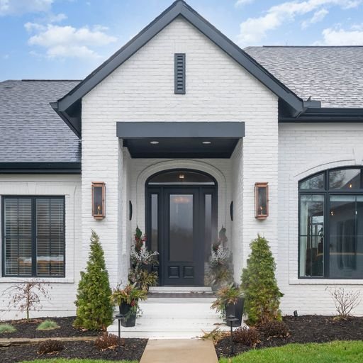 Front view of a modern white brick house with gray roofing, black front door, and large windows, with a small garden and pathway leading to the entrance.