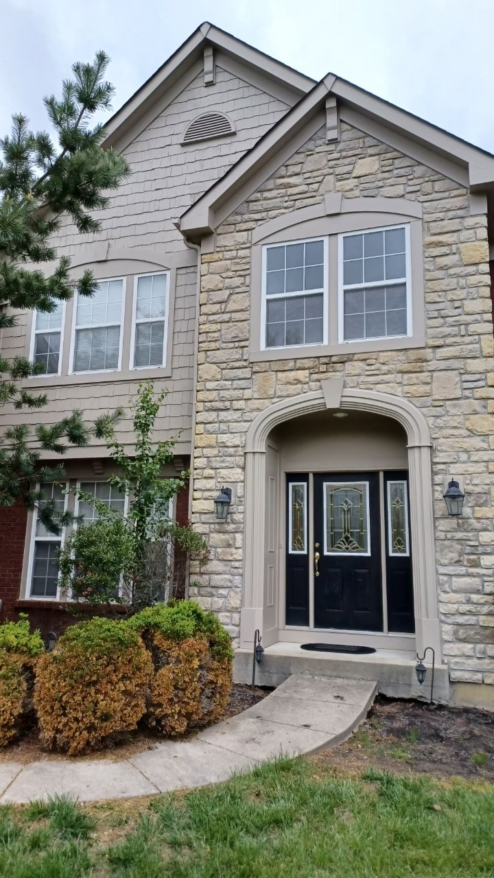 Front porch of a two-story house with stone and siding exterior, black front door with decorative glass, small walkway, and landscaped bushes.