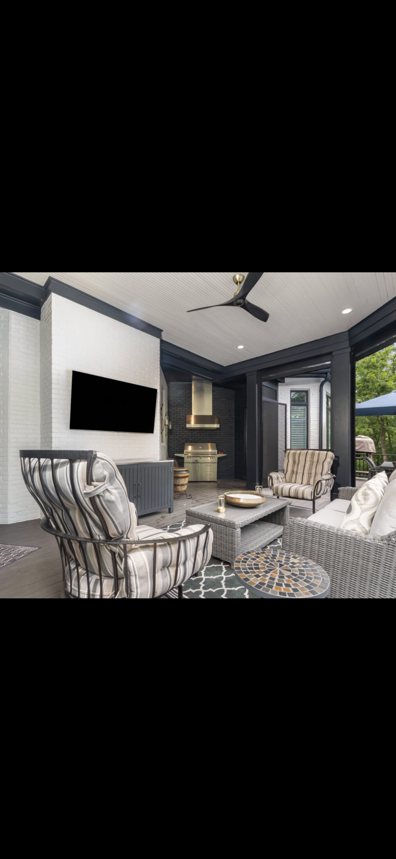 Covered porch with seating area, striped outdoor chairs, wicker sofa with cushions, a black ceiling fan, wall-mounted TV, and a ceiling with recessed lighting, overlooking a green outdoor space.