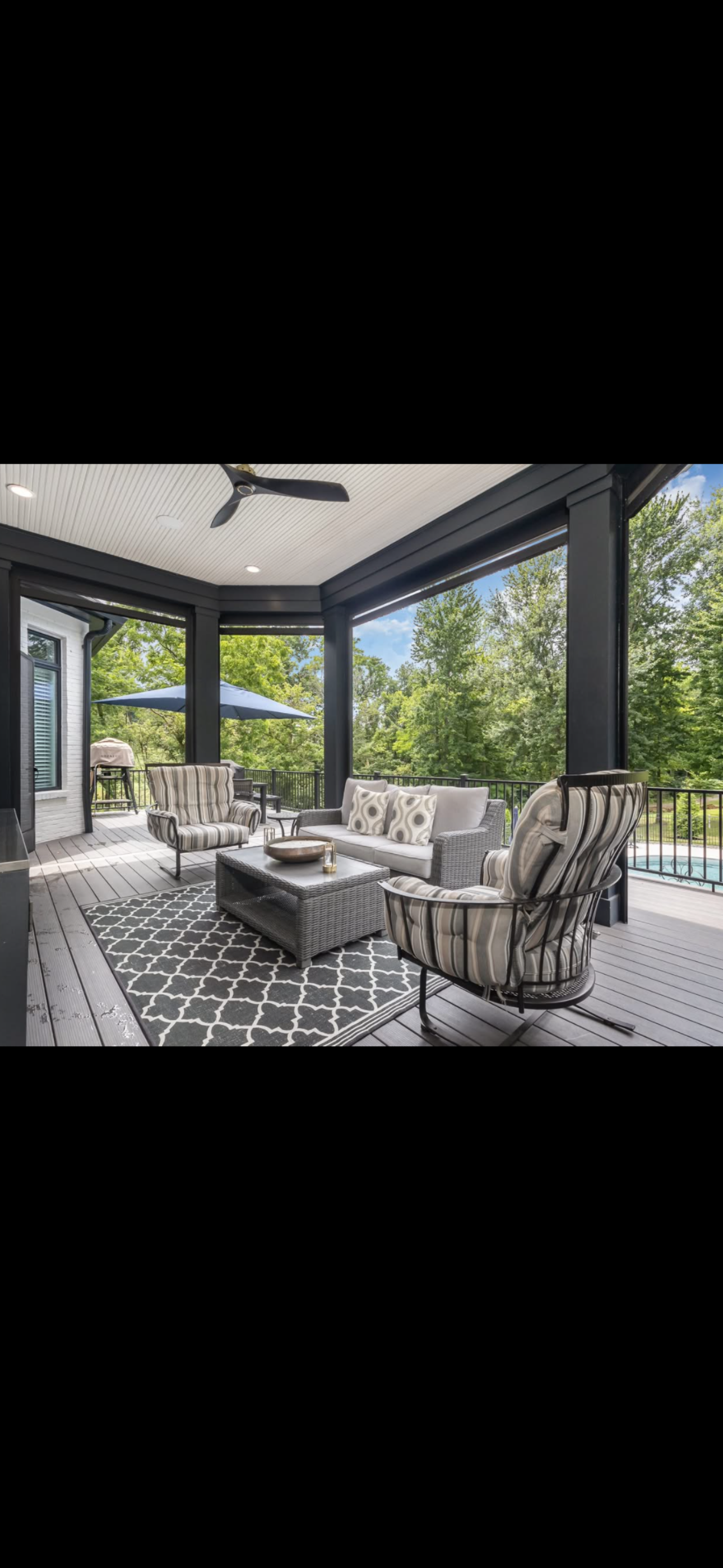 Covered outdoor porch with ceiling fan, furniture including striped armchair, sofa, and coffee table, black and white rug, umbrella, trees in the background.