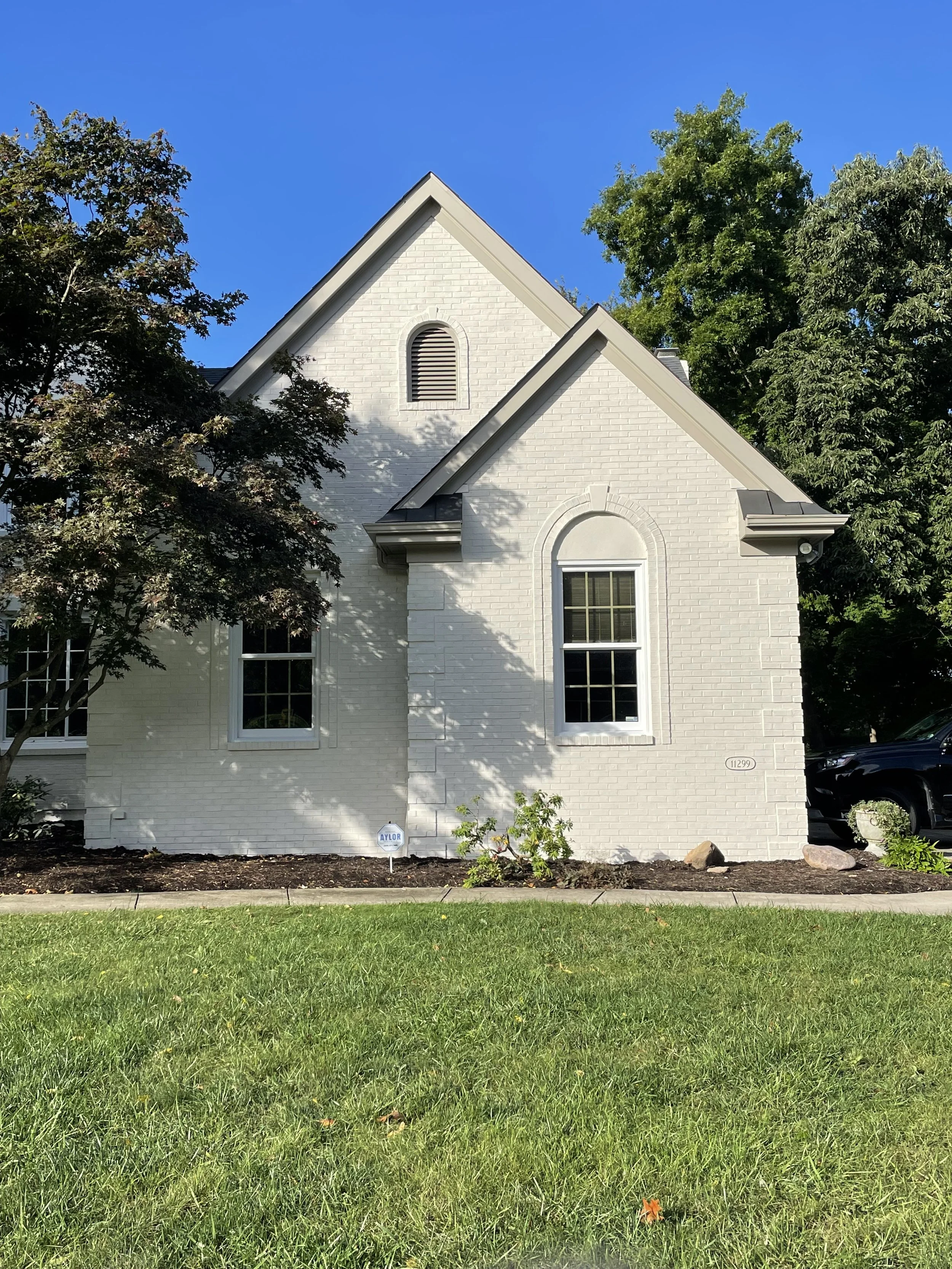Front view of a white brick house with three tall windows and a tall gabled roof, surrounded by trees and a grassy yard.