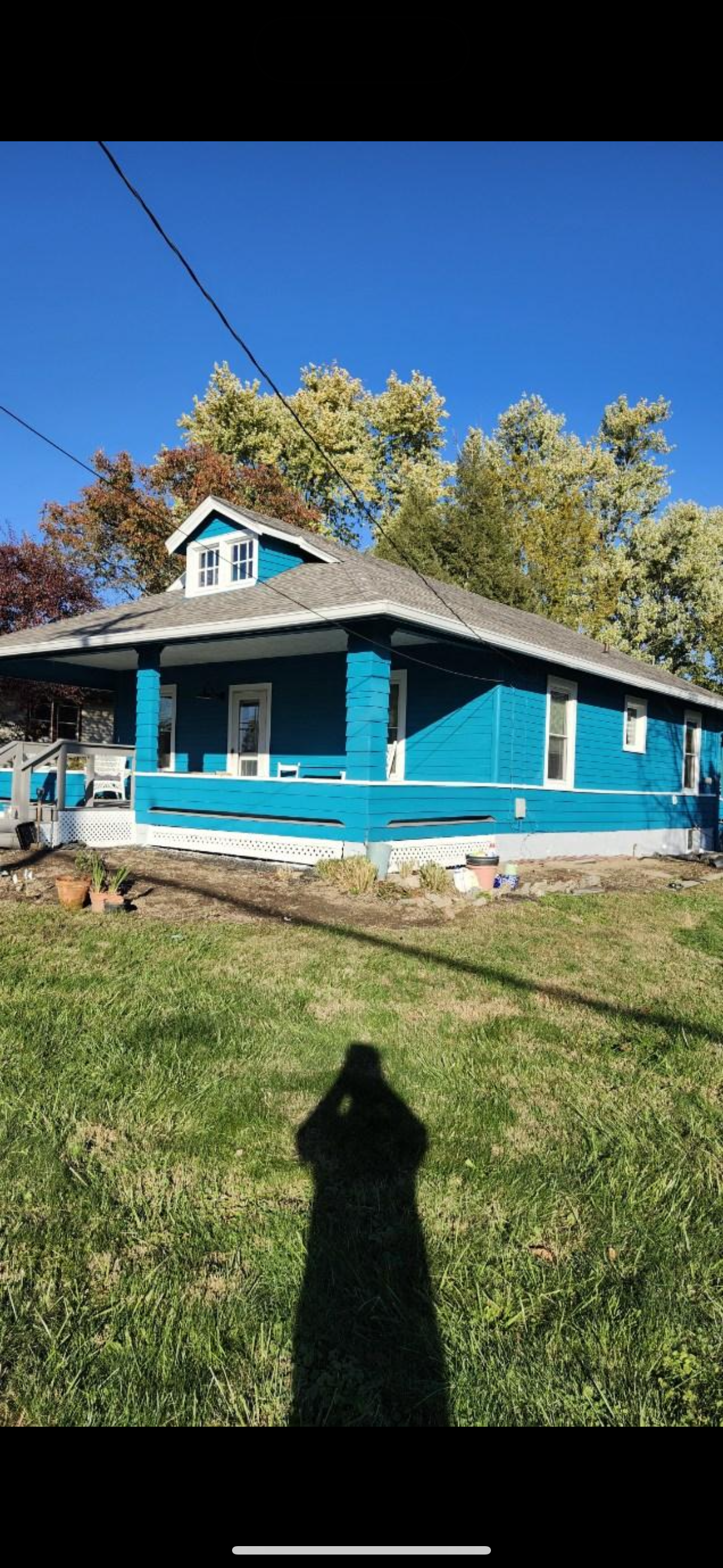 A blue house with a front porch and a small dormer window on the roof, set against a clear blue sky with green and autumn-colored trees in the background.