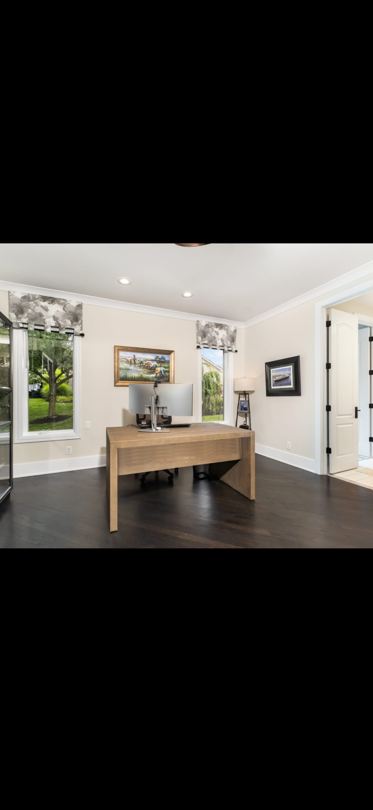 A home office with a wooden desk, dual monitors, and a computer mouse. There are large windows with patterned curtains, framed artwork on the walls, and a black wooden floor.