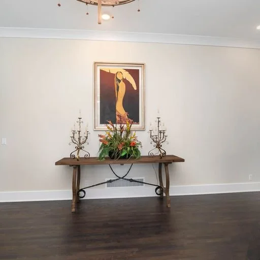 Empty room with a wooden table holding a floral arrangement and two candelabras, large framed artwork of a woman hanging on the white wall behind the table.