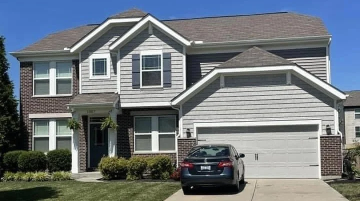 A two-story suburban house with gray siding and brick accents, a two-car garage, and a parked black car in the driveway, with a well-maintained lawn and bushes in front.