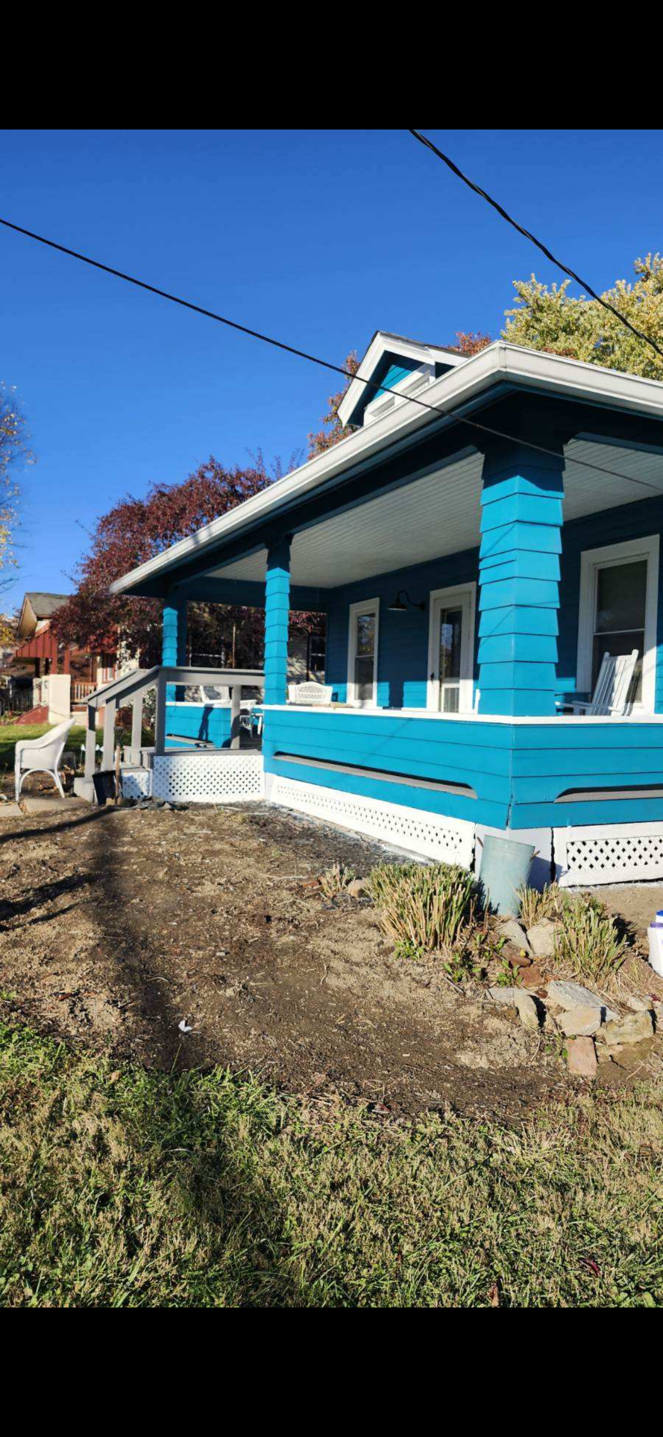 A blue house with a white porch railing, white trim, and a small front yard with dirt and some plants, under a clear blue sky.