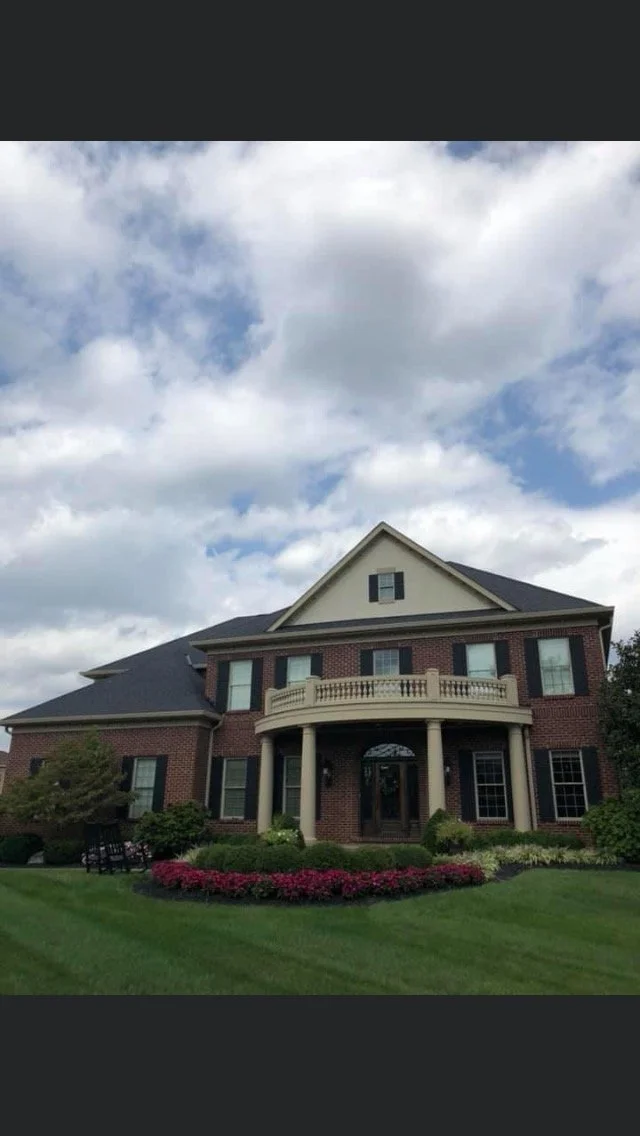 A large brick house with a front porch, balcony, and multiple windows, surrounded by a green lawn and flower beds, under a partly cloudy sky.