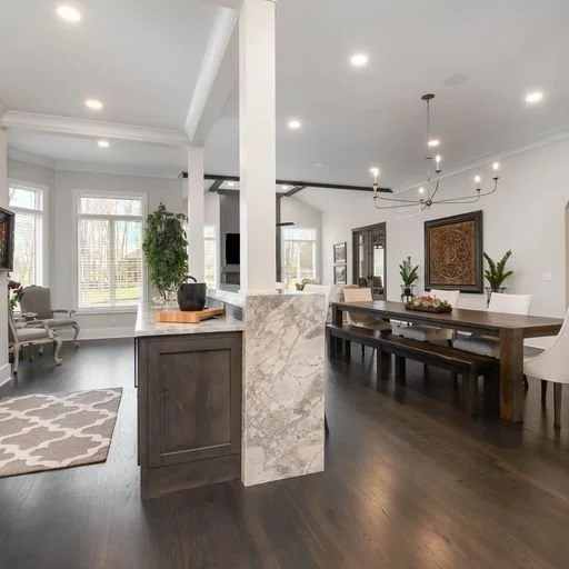 Open-concept living and dining area with white walls, dark wood flooring, and large windows, featuring a gray armchair, potted plants, and a wooden dining table with a chandelier above.