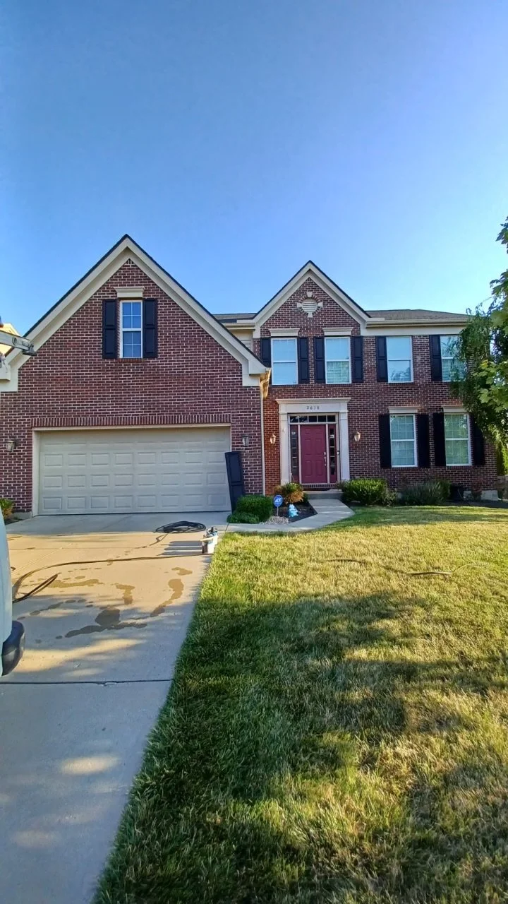 A two-story brick house with a red door, black shutters, and a garage, surrounded by a manicured lawn and a small garden area.