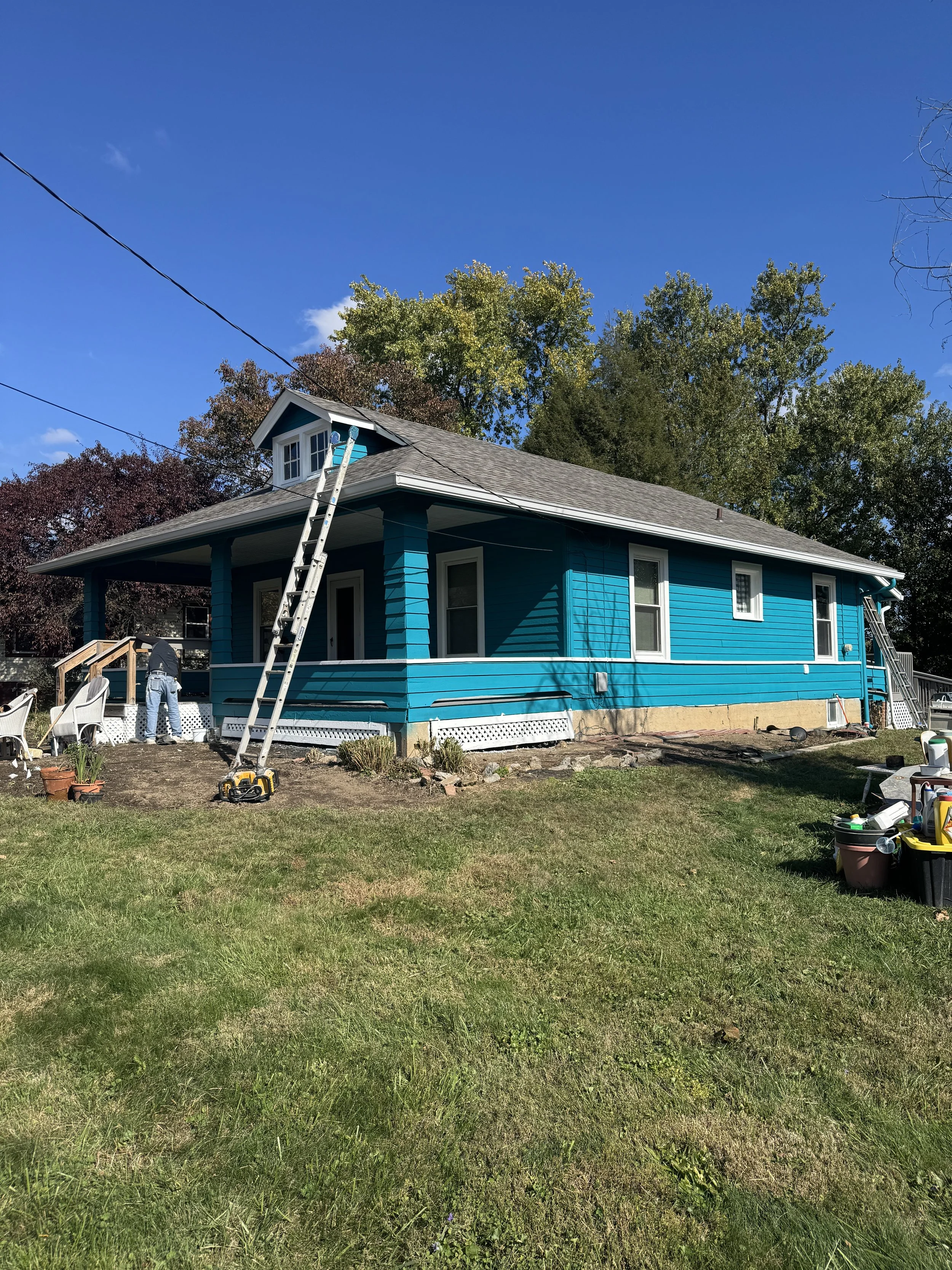 A bright blue house with a covered porch and a dormer window on a roof. There are ladders leaning against the house and some tools and equipment on the ground. The yard has a patch of dirt with some plants, and the background features trees with gree