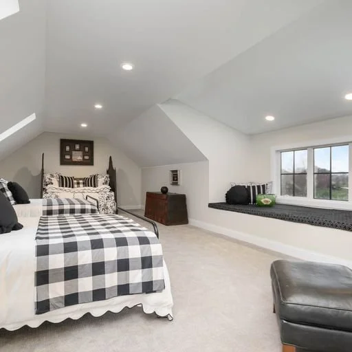 A spacious bedroom with two beds featuring black and white checkered bedding, a window seat with green pillows, and a window overlooking a rural landscape.
