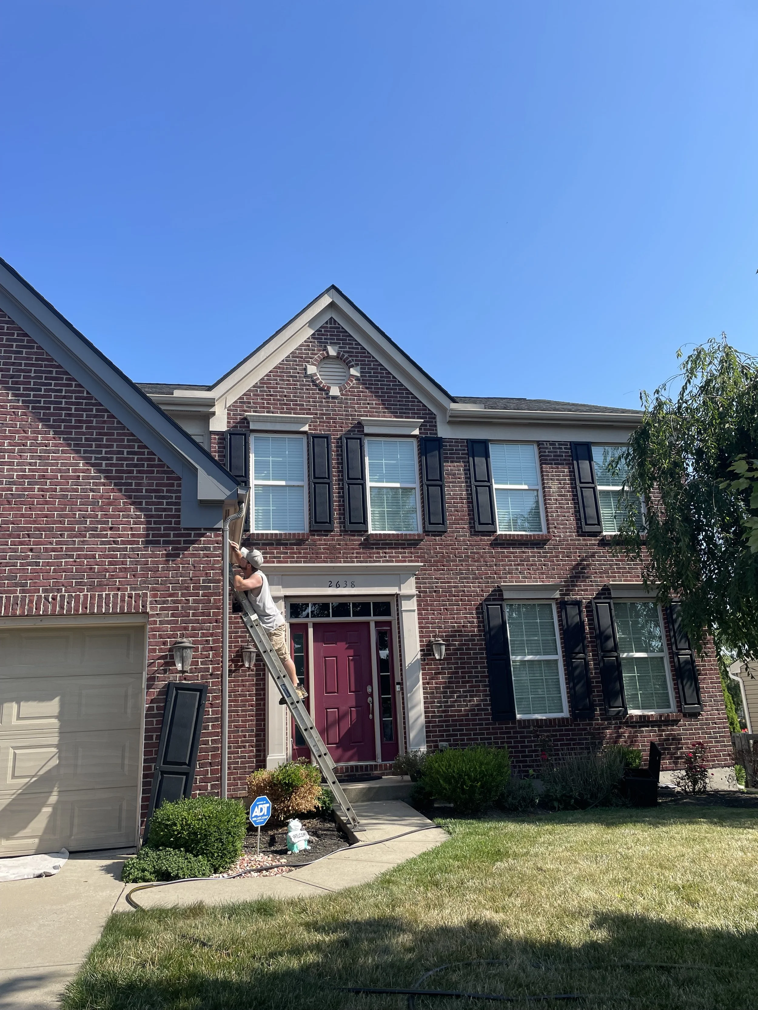 A person standing on a ladder cleaning gutters on a two-story brick house with black shutters. The house has a pink front door and several windows. There is a landscaped area with bushes and a small statue near the walkway. The sky is clear and blue.