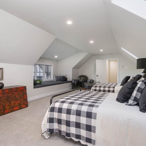 A bright attic bedroom with two beds, black and white checkered bedding, black pillows, a window seat, and a small red chest. The room has white walls and ceiling with recessed lighting.