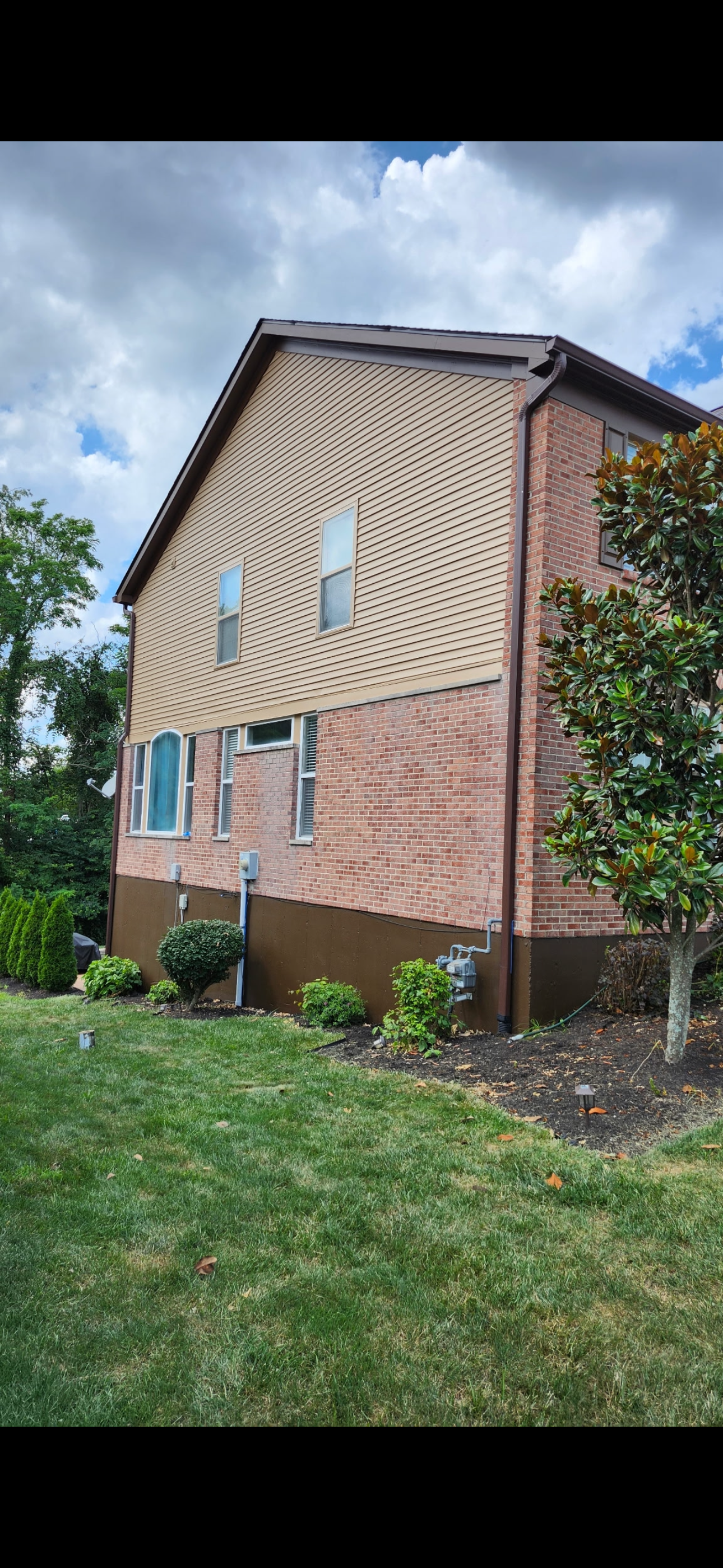 The image shows the side of a two-story house with brick and beige siding exterior. There are several windows, an arched window, and a small garden with bushes and a tree in the yard. The sky is partly cloudy.